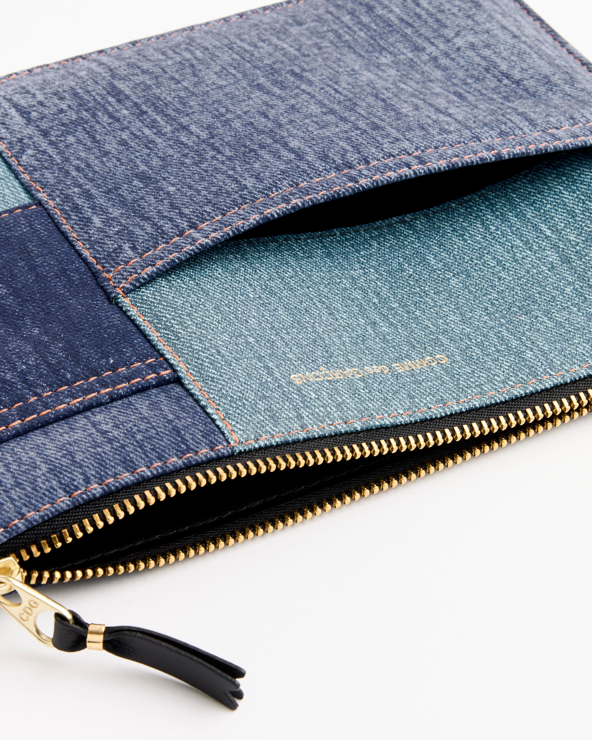 A close-up of the Comme des Garçons Wallet Denim Pocket Wallet in Blue, featuring patchwork denim, a gold zipper, visible stitching, and partially open to show the inner lining and gold embossed text on the pocket.