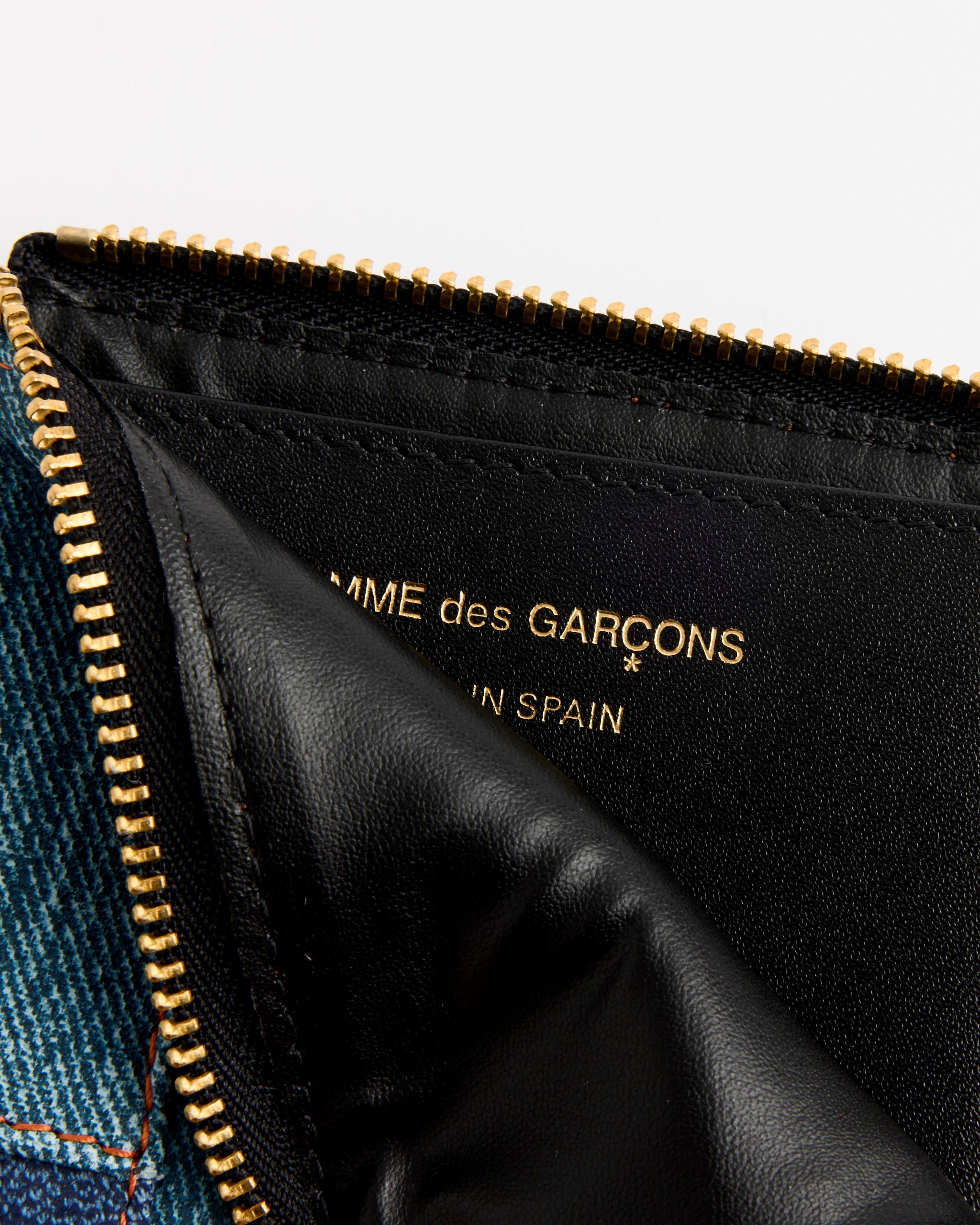Close-up of the Comme des Garcons Wallet Denim Pocket Wallet in Blue, featuring a gold zipper and partially open to show gold text inside; denim fabric on the left highlights its stylish design.