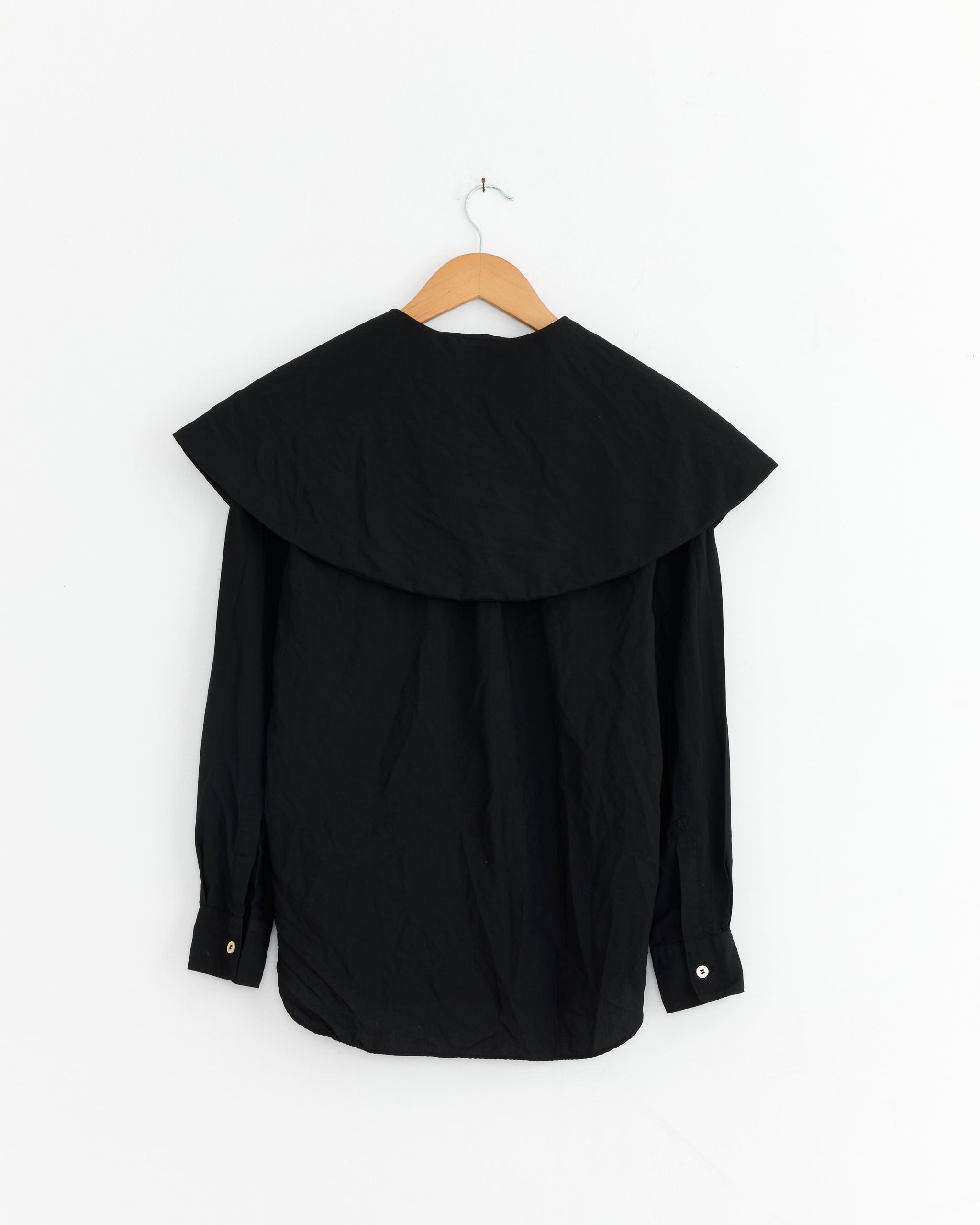 The Comme des Garçons Comme des Garçons B016 Blouse in Black, featuring a large wide collar, is crafted from Japanese polyester and shown from the back hanging on a wooden hanger against a plain white wall.