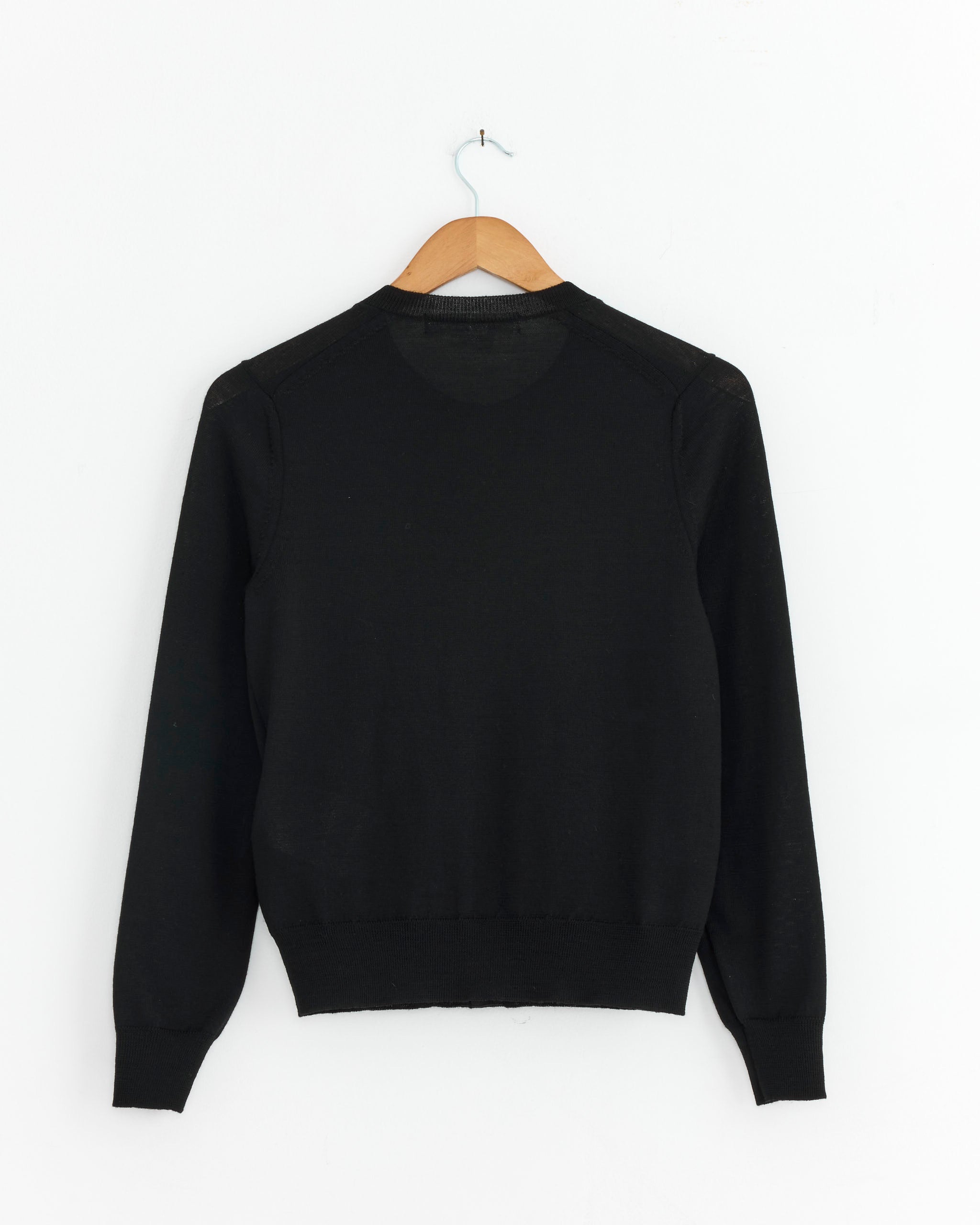 The N519 Cardigan in Black by Comme des Garçons Comme des Garçons, made from 100% wool, is displayed from the back on a wooden hanger against a white wall.