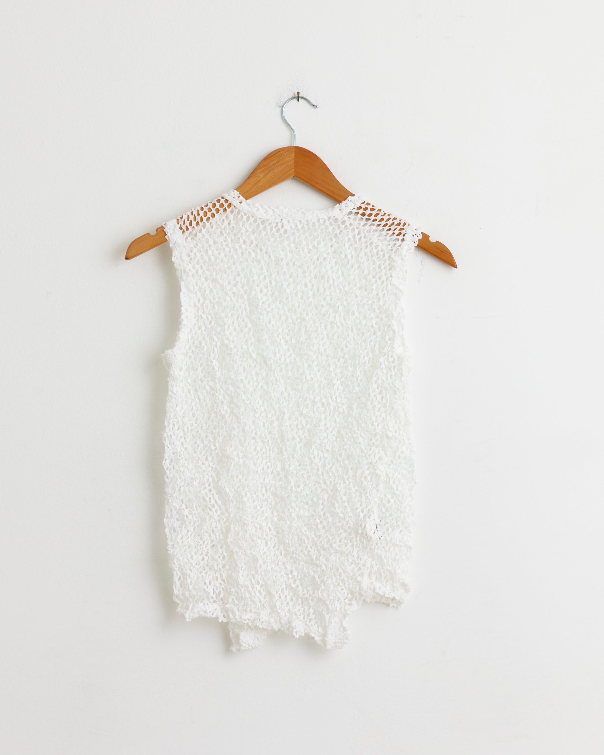 The T015 Top in White by Comme des Garçons Comme des Garçons is a sleeveless crochet design in 100% polyester, with airy weave and uneven hem. Displayed on a hanger against a white wall. Refer to size chart for the best fit.