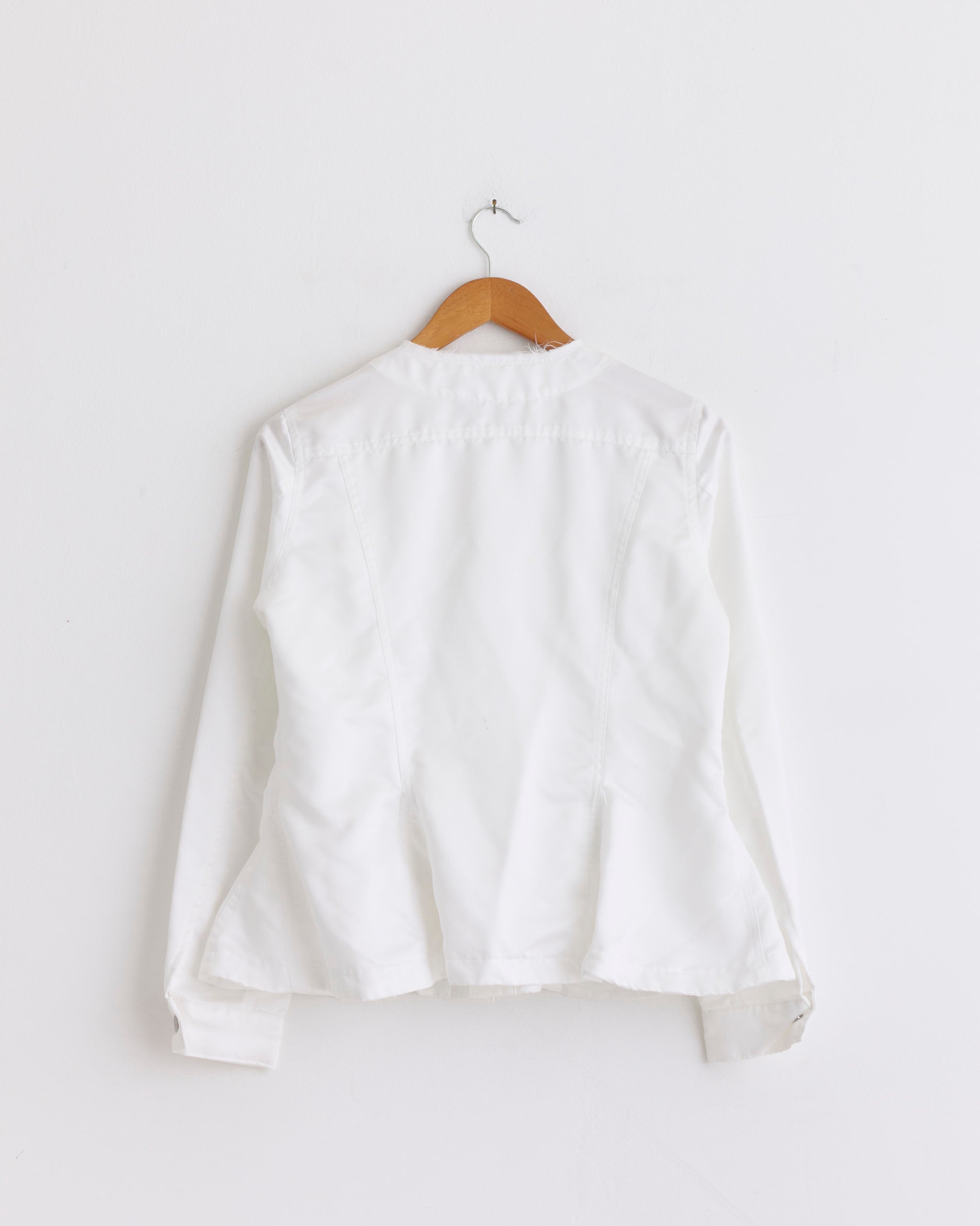 The J024 Jacket in White by Comme des Garçons Comme des Garçons hangs on a wooden hanger against a plain white wall, displayed from the back to highlight seam stitching details.