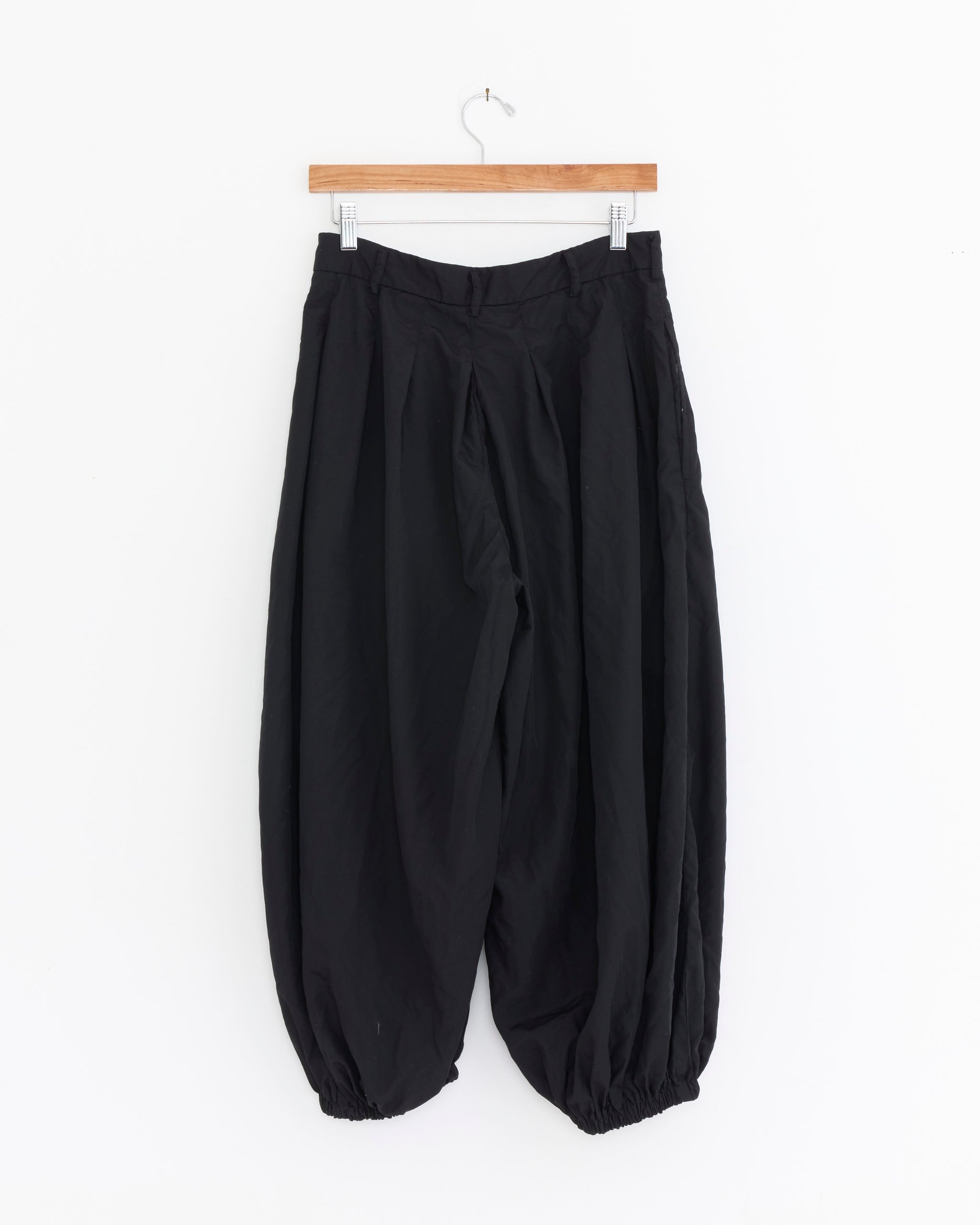 The P006 Pant in Black by Comme des Garçons Comme des Garçons features a loose fit with elastic waistband and ankle cuffs, shown hanging on a wooden hanger against a white background.