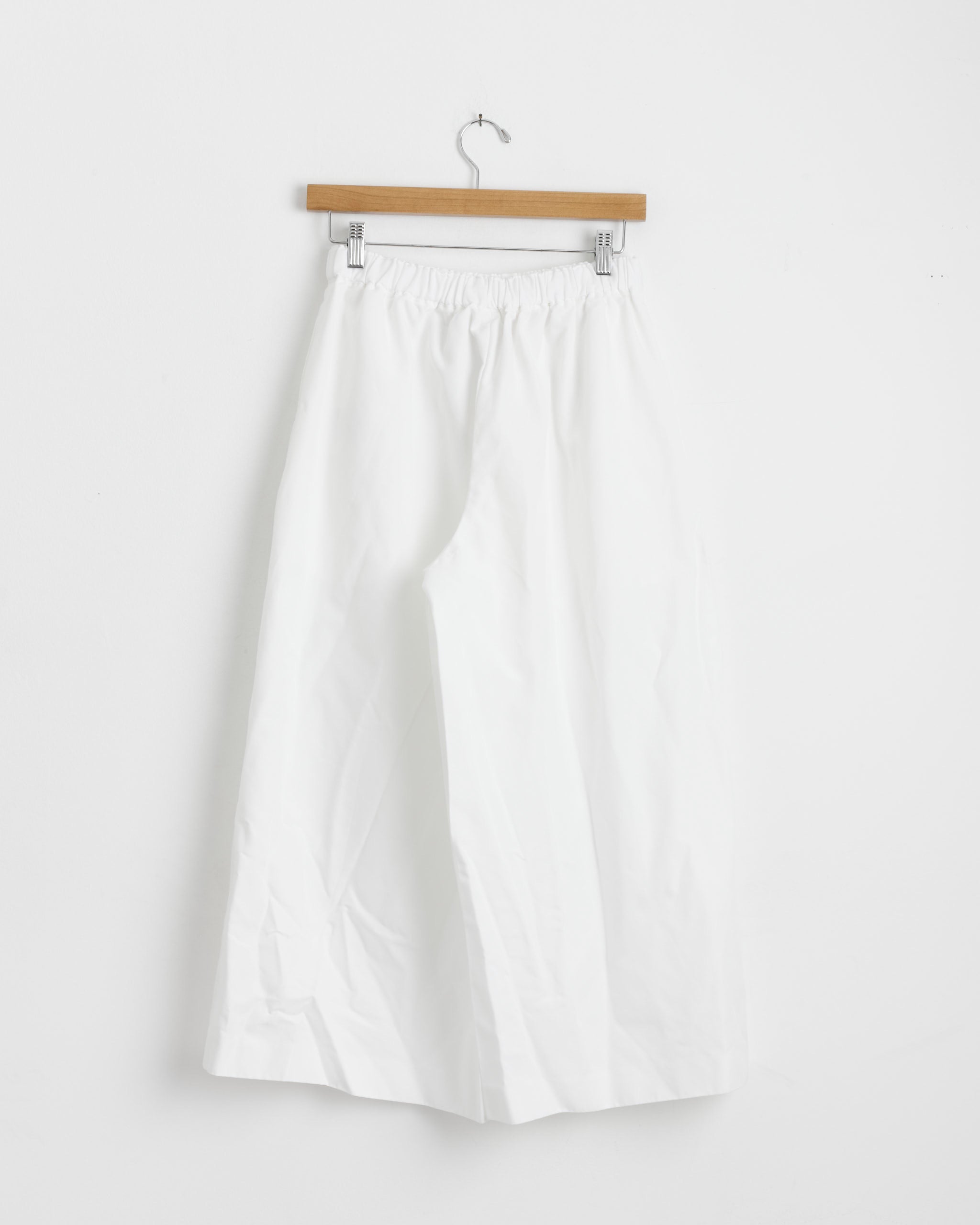 The Comme des Garçons Comme des Garçons P007 Pant in White, featuring a wide-leg cut and elastic waistband, hangs on a wooden hanger against a plain white wall.
