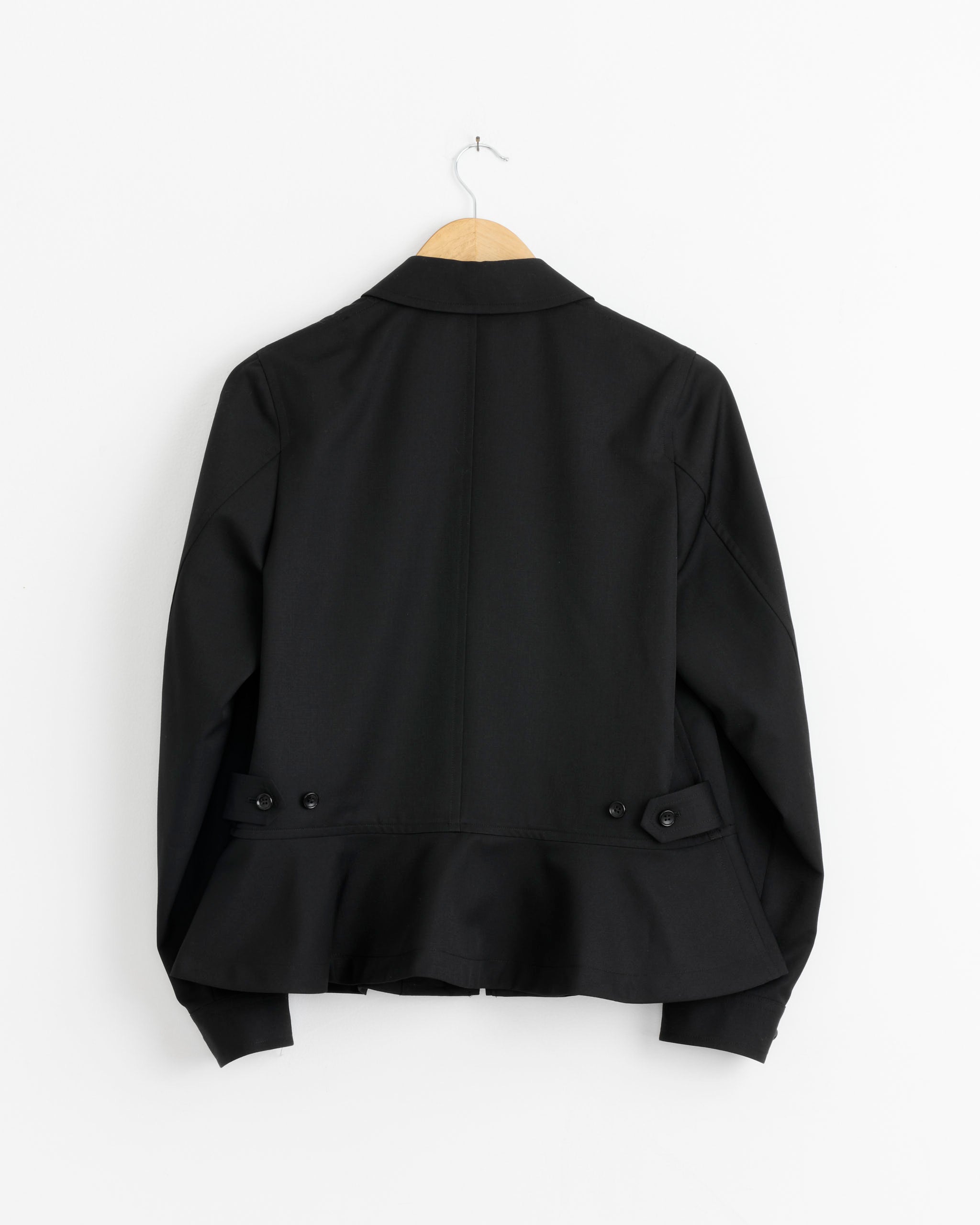 The J011 Jacket in Black by Comme des Garçons Comme des Garçons hangs on a wooden hanger against a white wall, displayed from the back with button details on both sides near the waist.