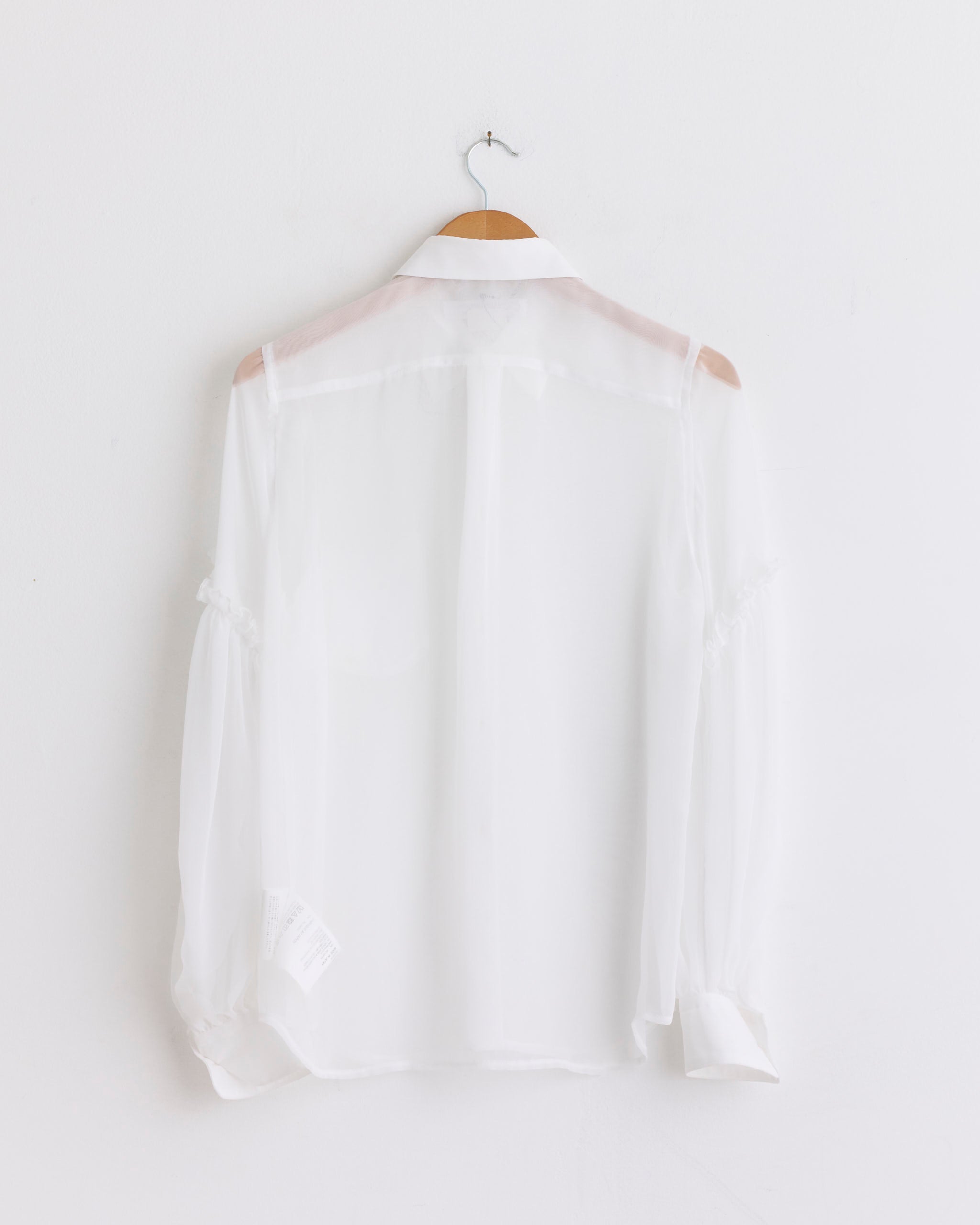 The RQ-B018 Blouse in Off White by Comme des Garçons Comme des Garçons is shown from the back, featuring sheer white long sleeves and displayed on a wooden hanger against a plain white wall.