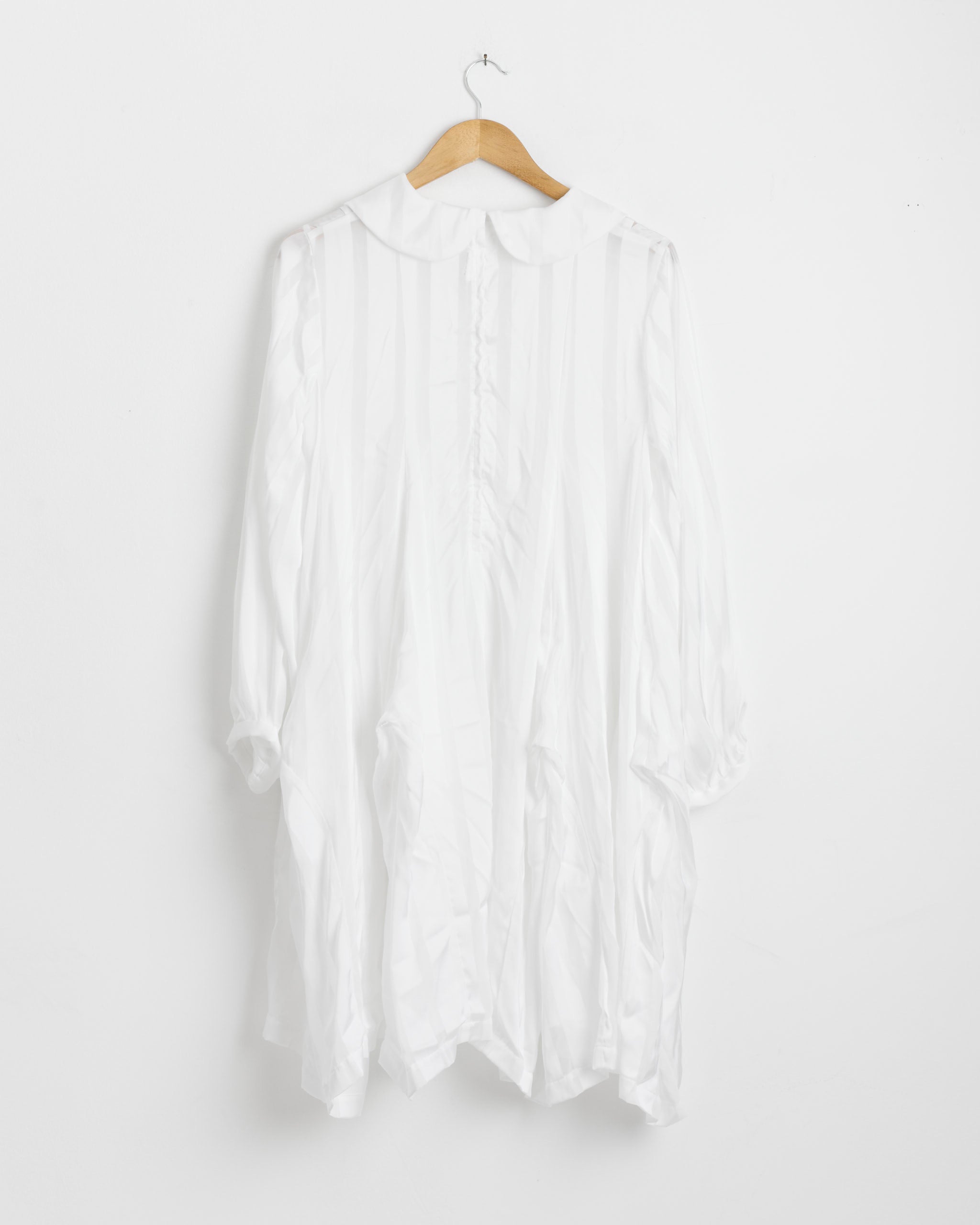 The B001 Shirt Dress in White by Comme des Garçons Comme des Garçons is a long-sleeved, loose-fitting dress with a Peter Pan collar and front buttons, shown on a wooden hanger against a white wall.