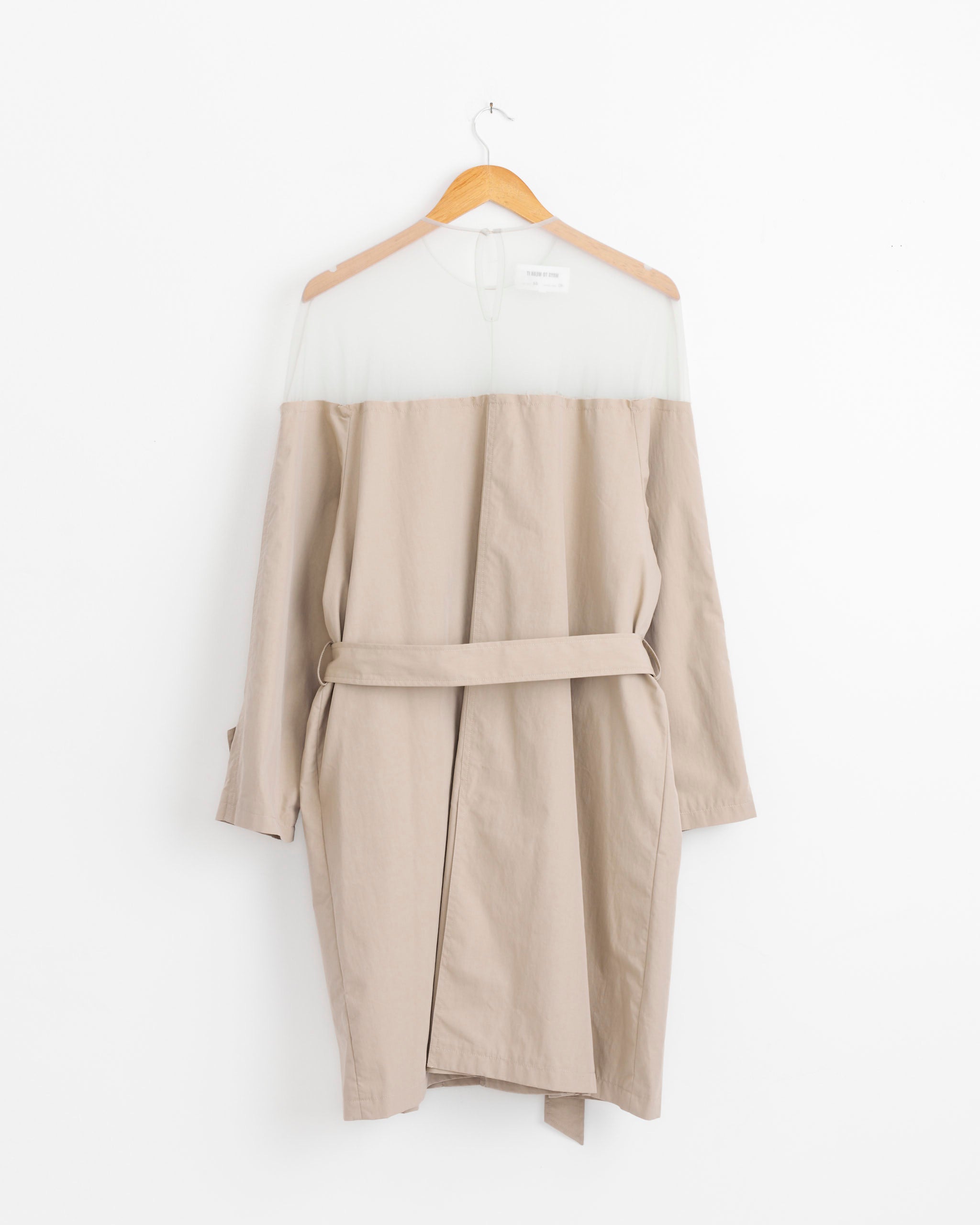 The Illusion Midi Trench Dress in beige by MM6 Maison Margiela hangs on a wooden hanger against a white wall, featuring long sleeves, a matching belt, and a minimalist design.