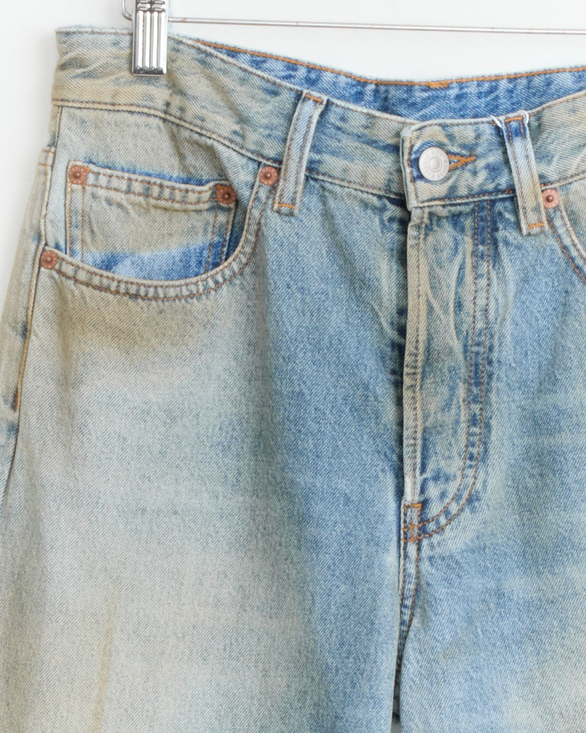 Close-up of MM6 Maison Margiela 5 Pocket Pant in Medium Blue hanging on a hanger, highlighting the waistband, button, front pockets, and belt loops.