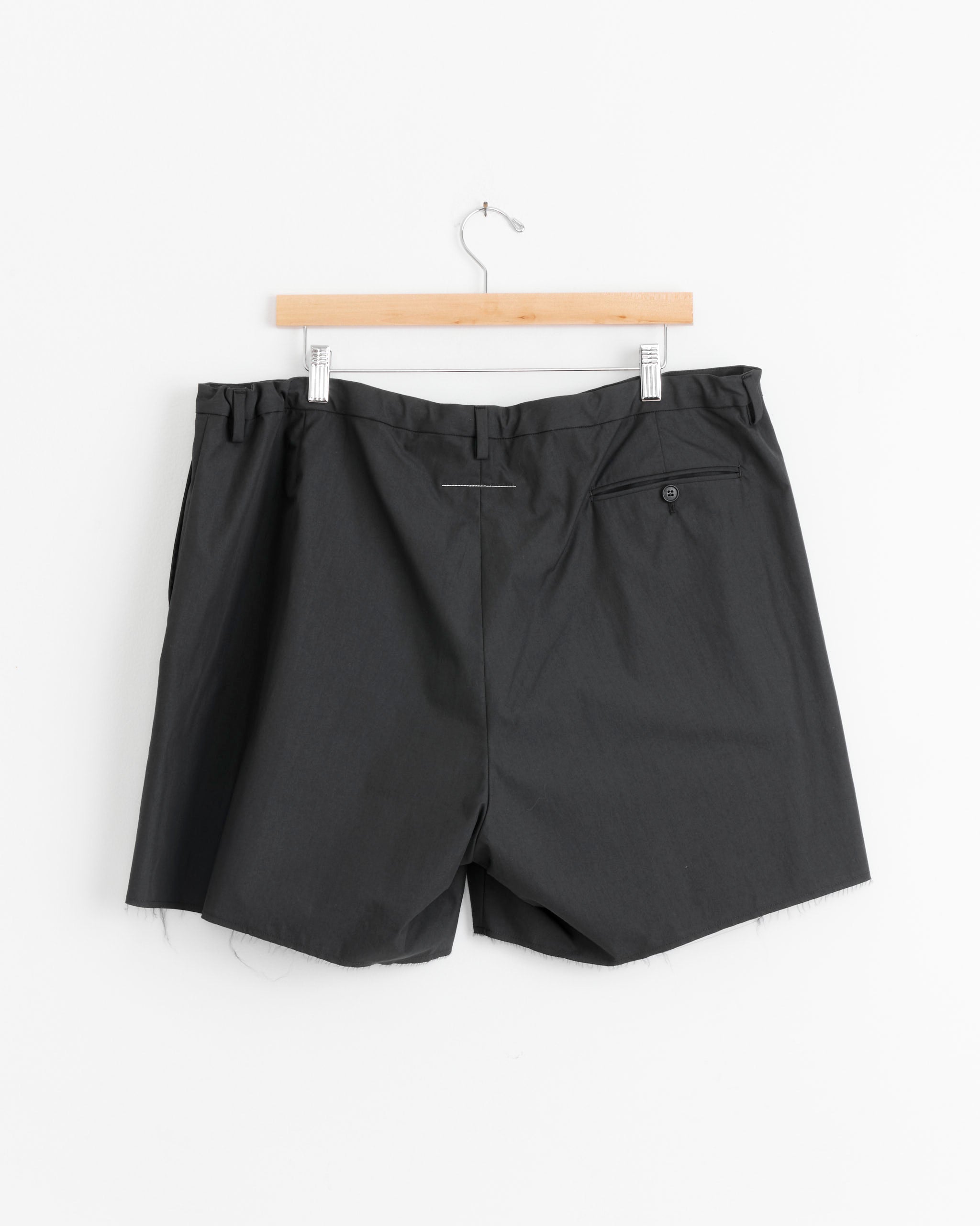 The MM6 Maison Margiela Frayed Short in Anthracite Grey features an elastic waistband, a single buttoned back pocket, and frayed hems. Displayed on a wooden hanger against a white background.