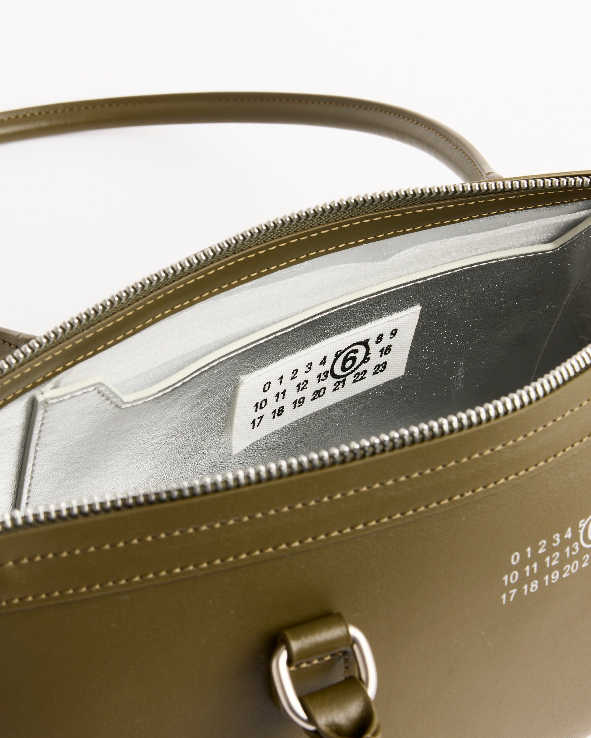 A close-up of the MM6 Maison Margiela New Bauletto J Medium Bag in khaki shows its leather exterior with signature numeric artwork, a silver zipper, and an interior white label featuring black numbers.