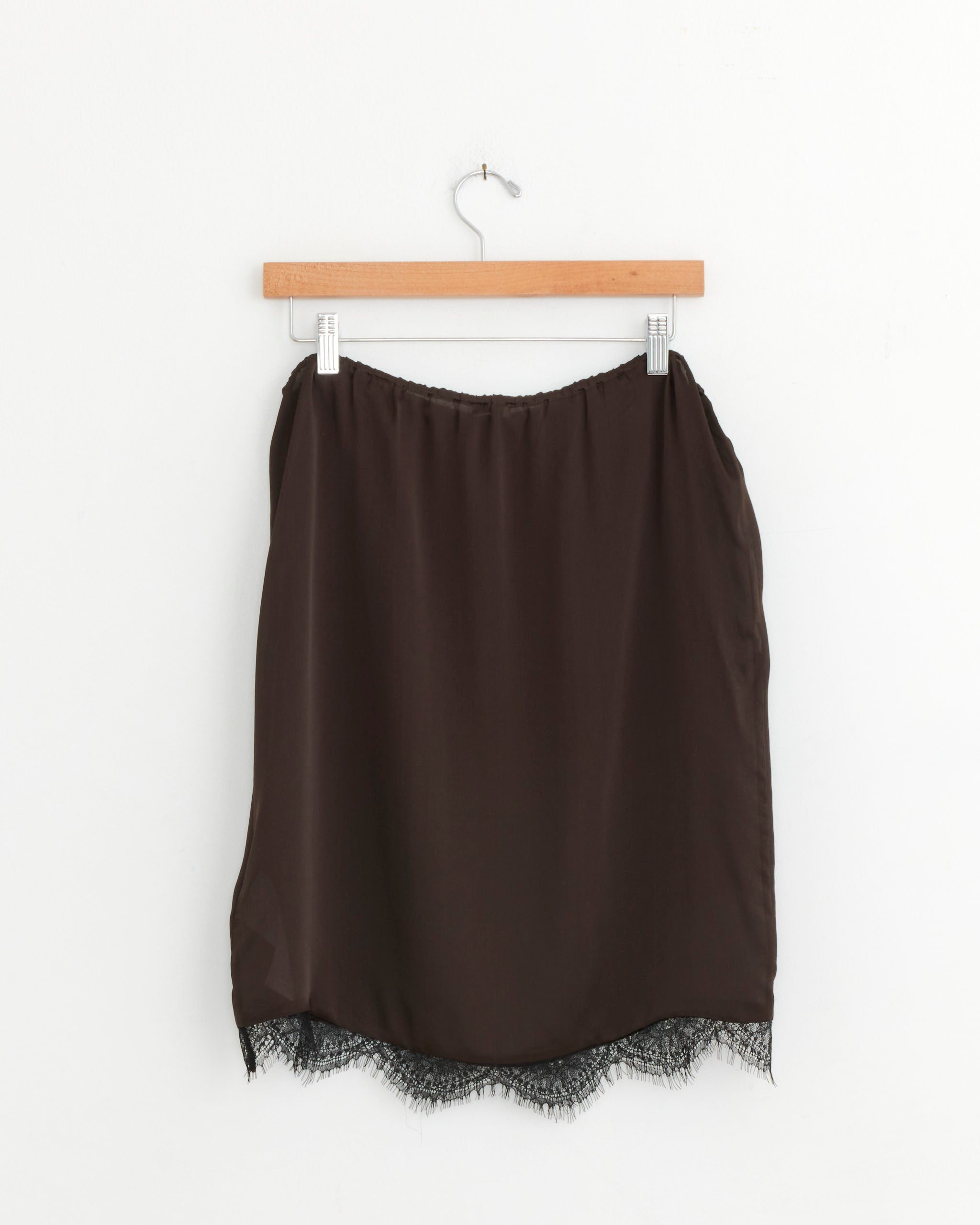 A bark-colored satin ST. AGNI Fine Lace Skirt with a delicate lace-trimmed hem hangs on a wooden hanger against a plain white wall.