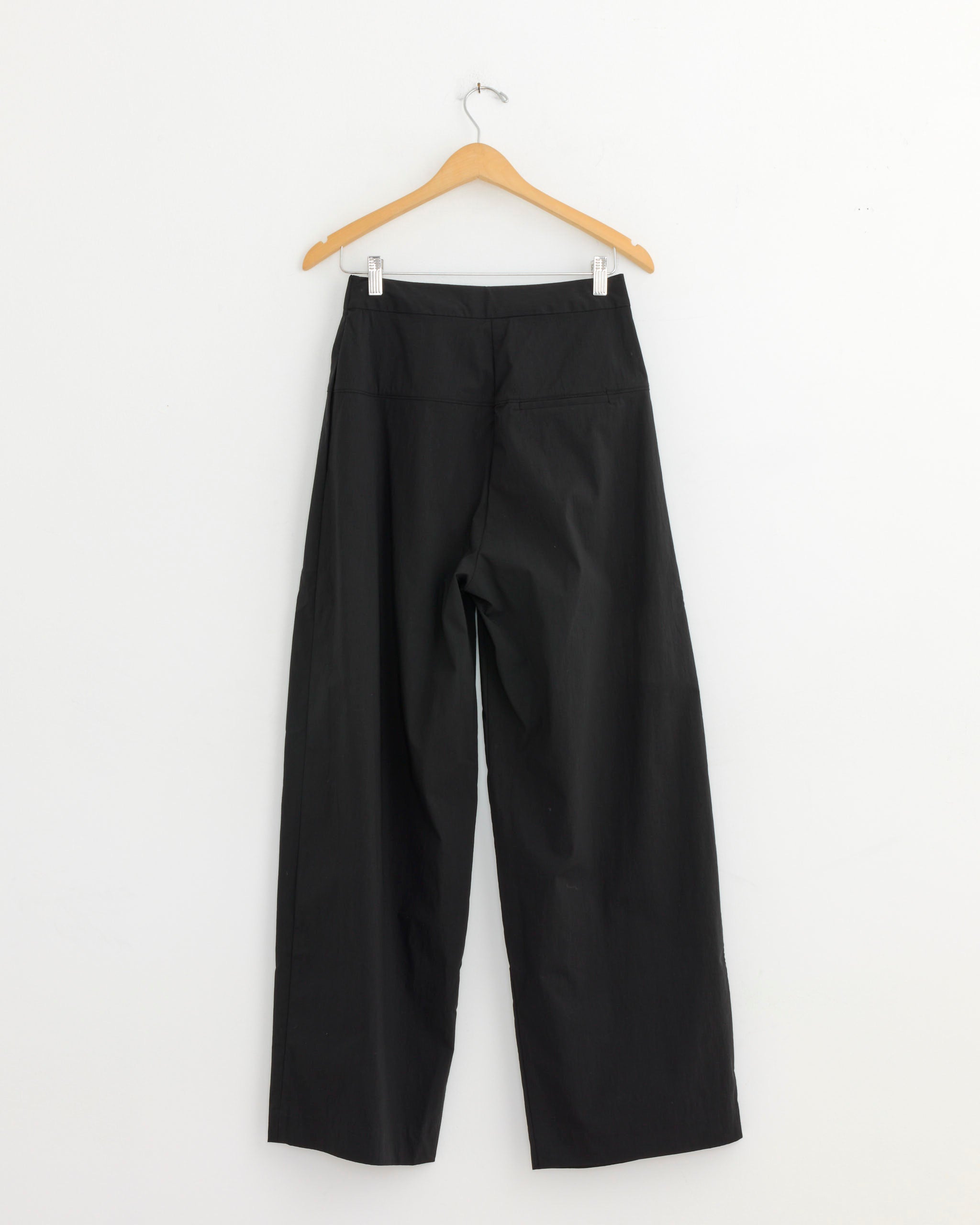 The ST. AGNI Relaxed Pleat Pant in Black, featuring a mid-rise waist and tailored finish, hangs on a wooden hanger against a plain white wall.