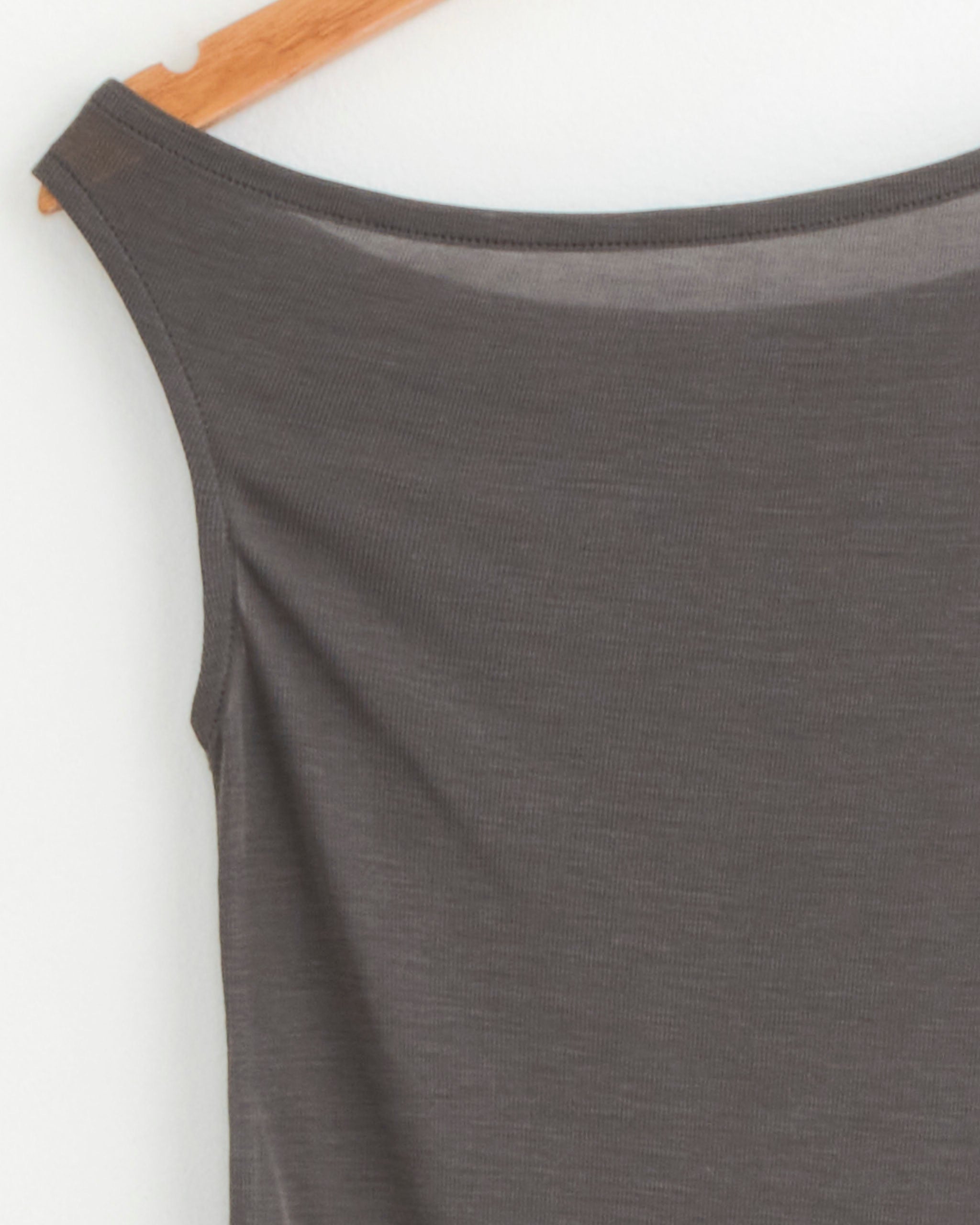 A close-up of the ST. AGNI Fine Jersey Asymmetrical Top in Soot on a wooden hanger against a white wall.