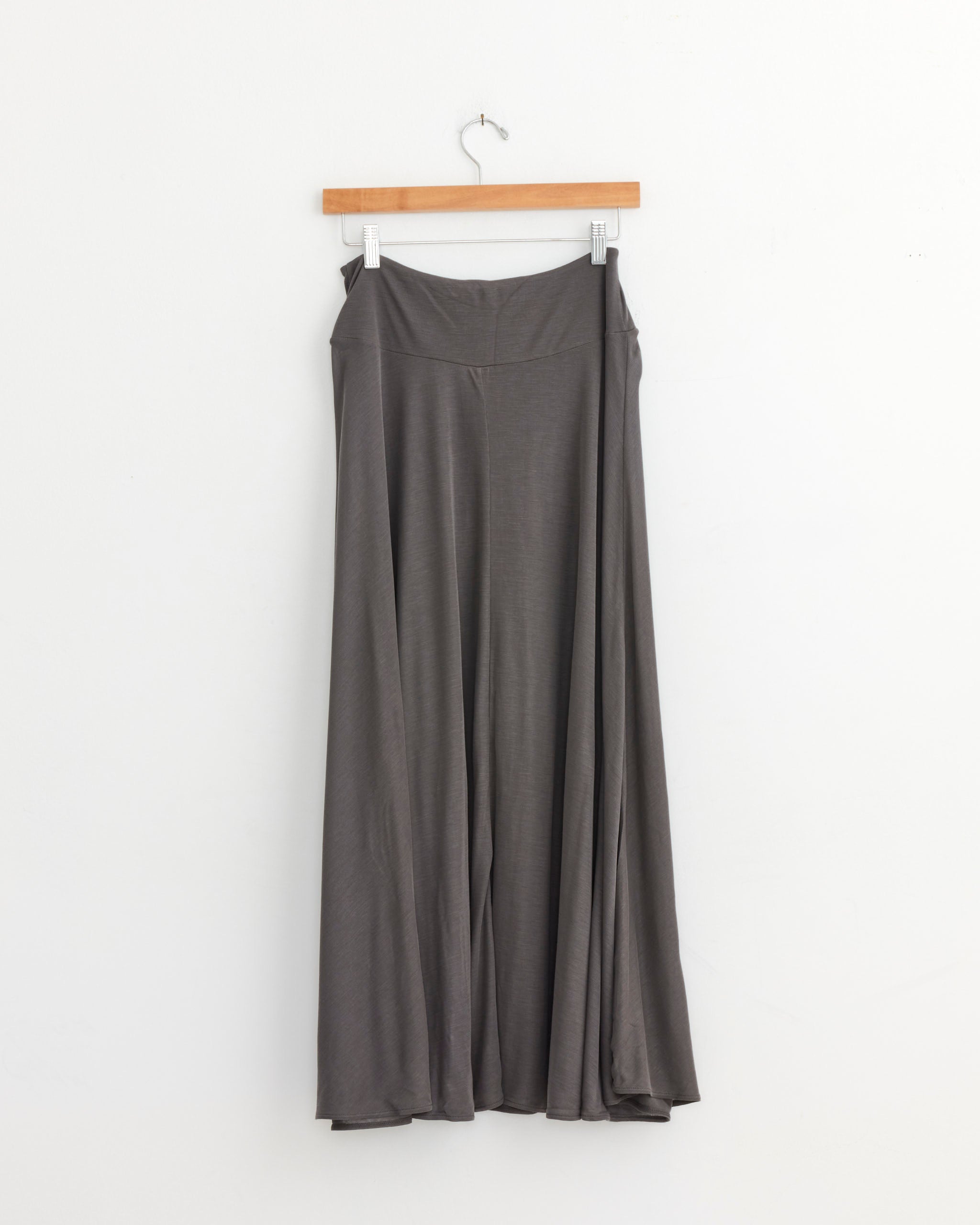 The ST. AGNI Fluid Jersey Skirt in Soot hangs on a wooden hanger against a plain white wall, highlighting its elegant and flowy silhouette.