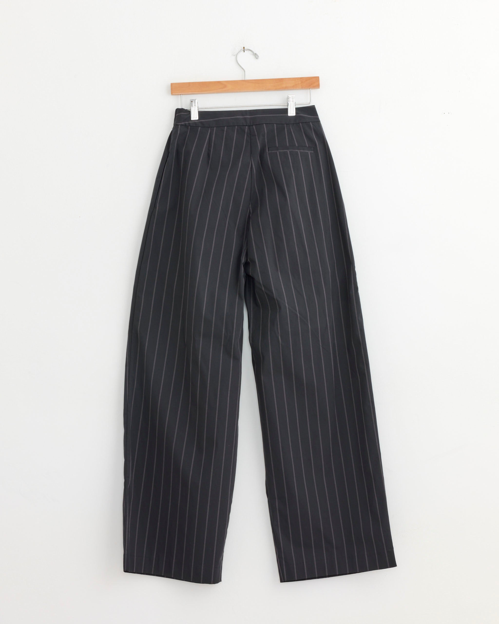 The ST. AGNI Relaxed Pleat Pant in Ink Stripe, featuring white vertical pinstripes, a mid-rise waist, straight-leg fit, tailored finish, and single back pocket, hangs on a wooden hanger against a plain white wall.