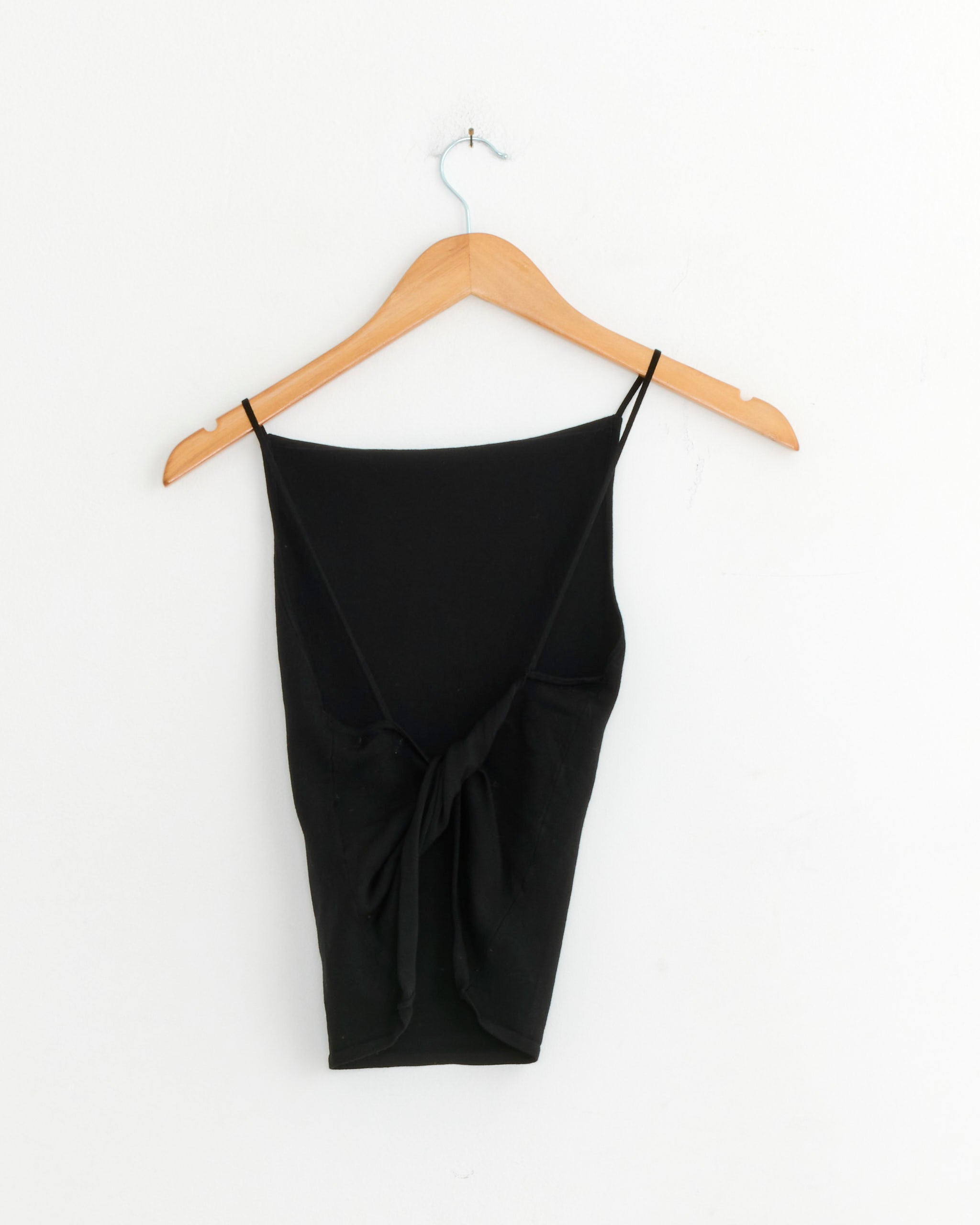 The ST. AGNI Twist Back Knit Top in Black, a sleeveless crop top with thin straps and a twist detail, is displayed on a wooden hanger against a plain white wall.