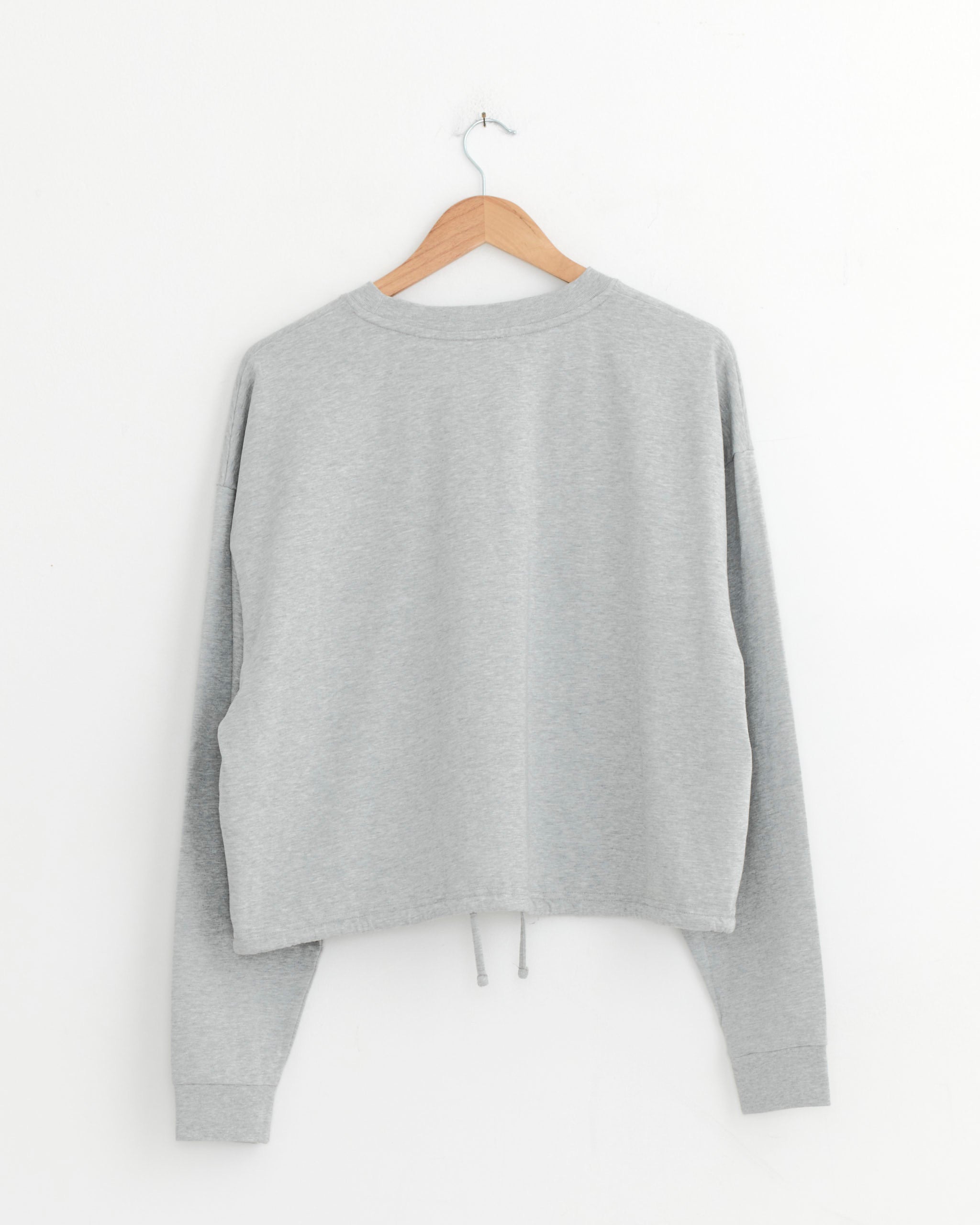 The ST. AGNI Drawstring Long Sleeve Top in Light Grey Marle, crafted from 100% cotton, is shown hanging on a wooden hanger against a white wall, displayed from the back.