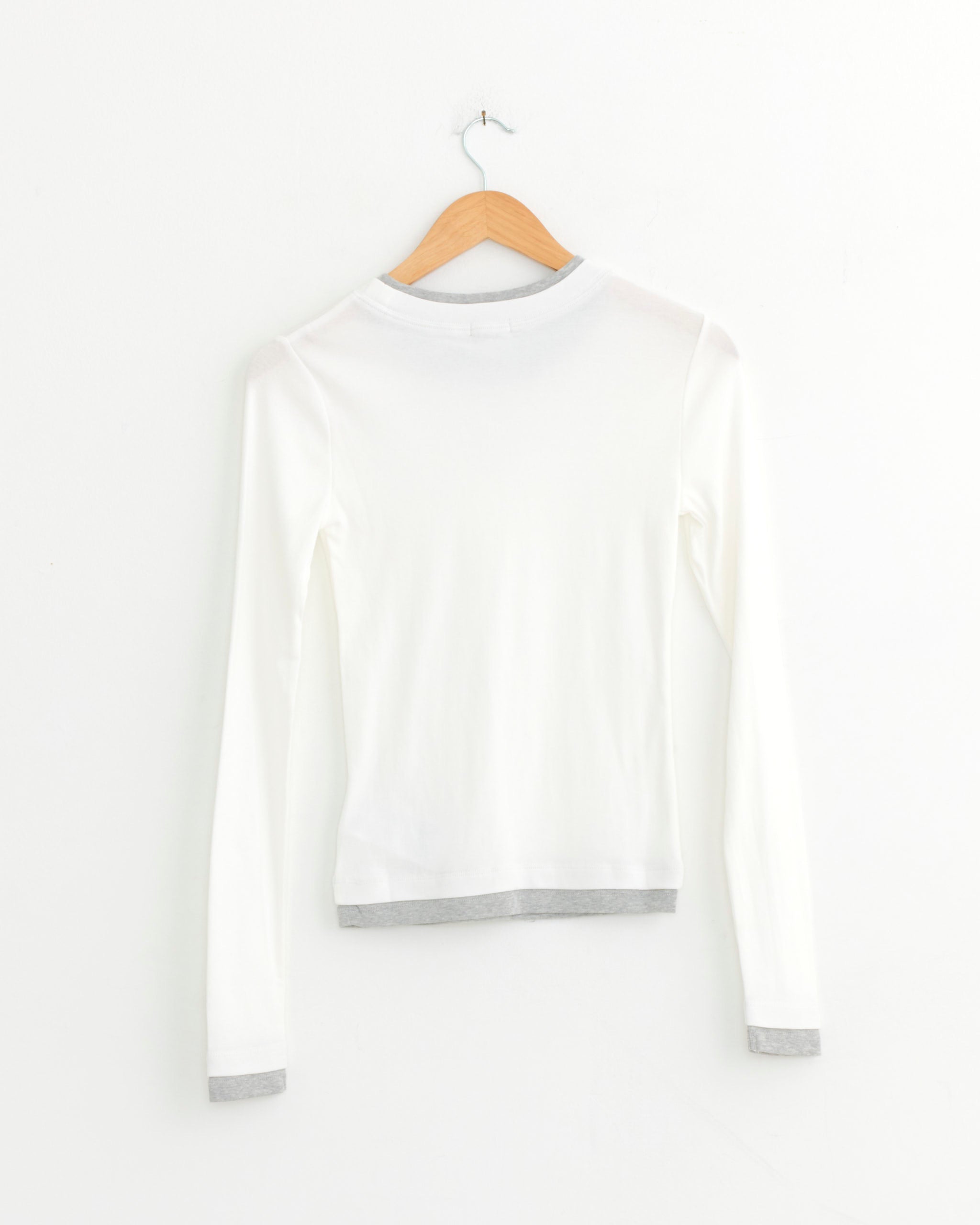 The ST. AGNI Layered Long Sleeve Top in White, featuring gray trim on the collar, cuffs, and hem, is displayed from the back on a wooden hanger against a white wall—an everyday essential silhouette.