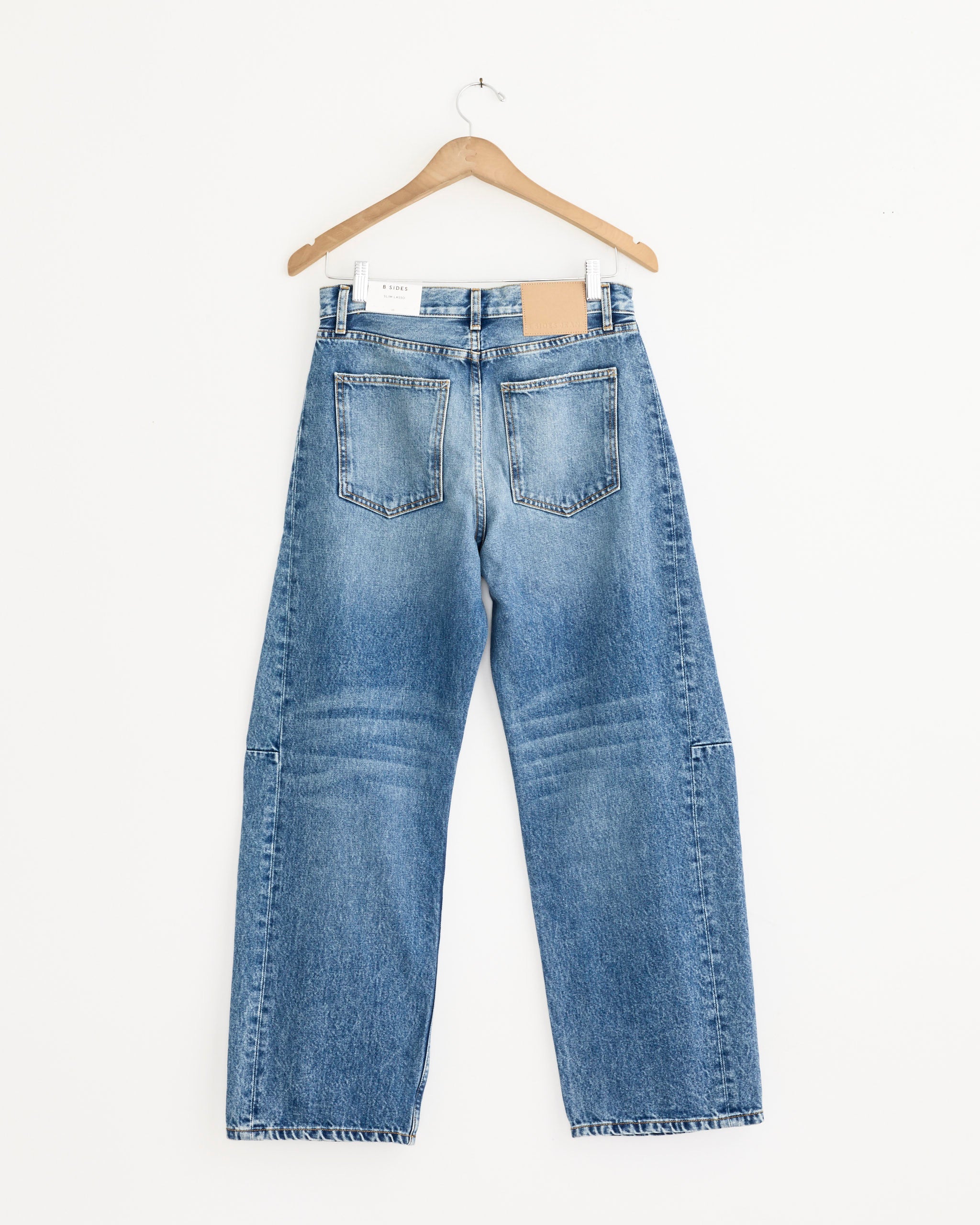 The B Sides Slim Lasso in Jane Wash is a pair of blue cotton denim jeans with a straight leg, shown from the back on a wooden hanger. Made in the USA, they feature two back pockets and a brown leather patch on the waistband.