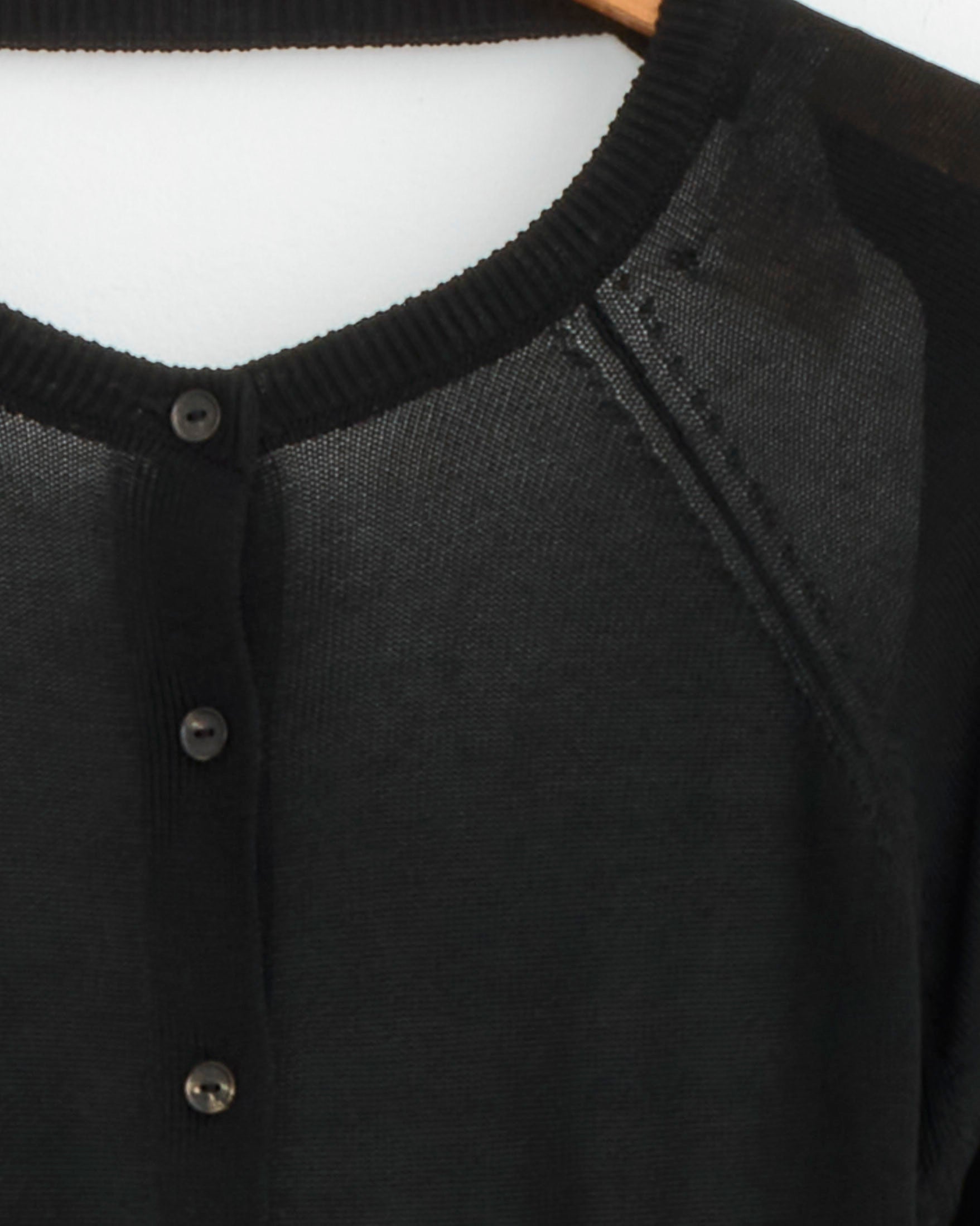 Close-up of the upper part of CORDERA’s Open Back Cardigan in Black on a wooden hanger, highlighting its sheer fabric, ribbed neckline trim, and three visible black buttons.