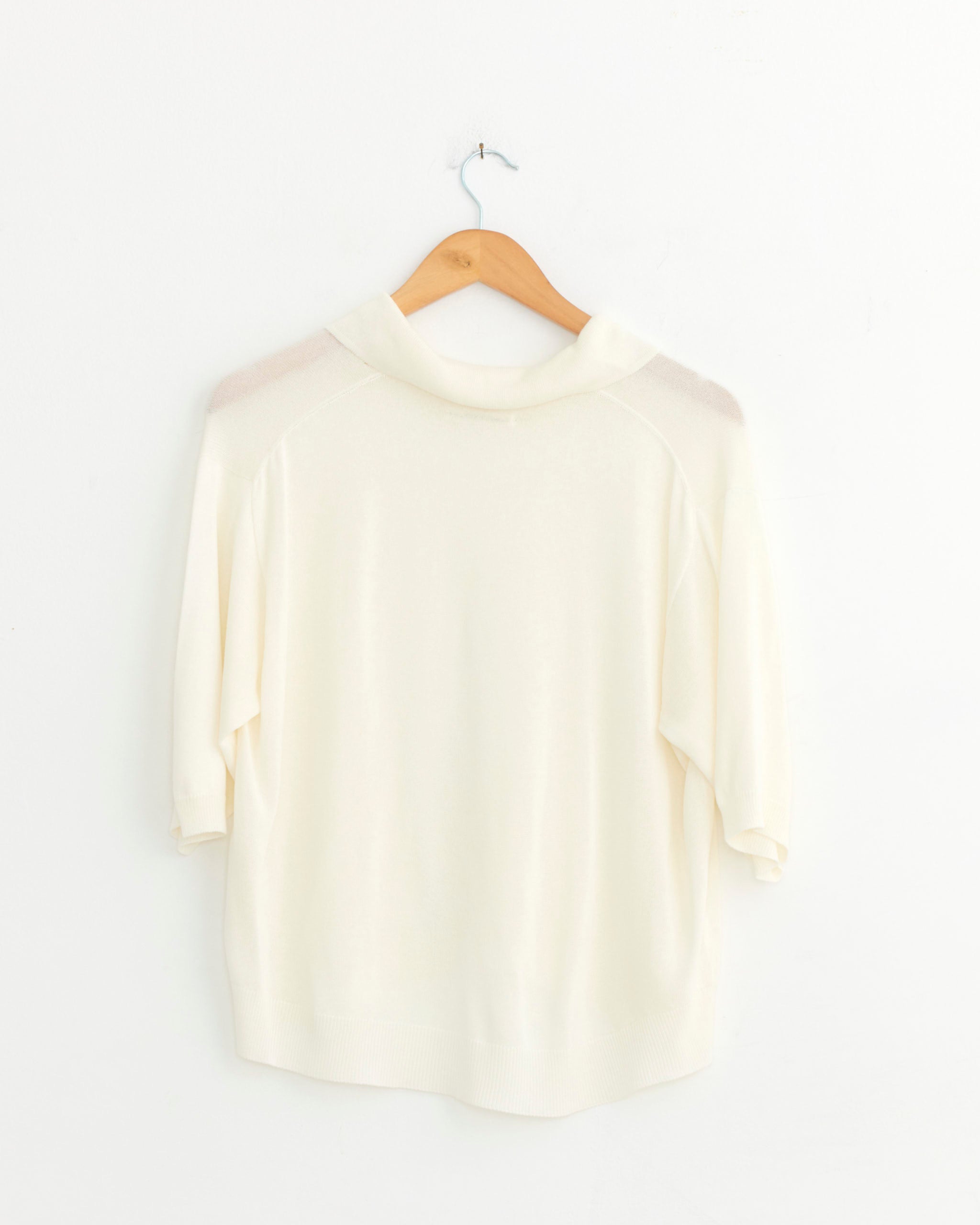 The CORDERA Lace Polo in Garza, a super-light white knit short-sleeved shirt with a collar, is shown hanging on a wooden hanger against a plain white wall with the back facing forward.