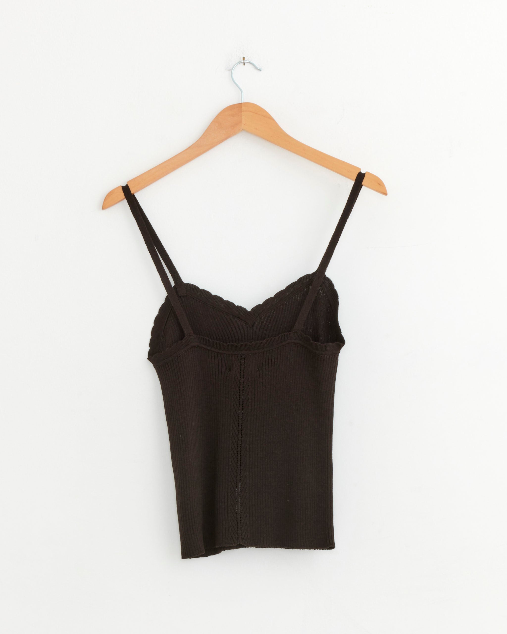 A brown CORDERA Ribbed Tank with thin straps and a scalloped neckline hangs on a wooden hanger against a plain white wall. Expertly crafted in Spain, this silk-cotton blend tank offers effortless style.