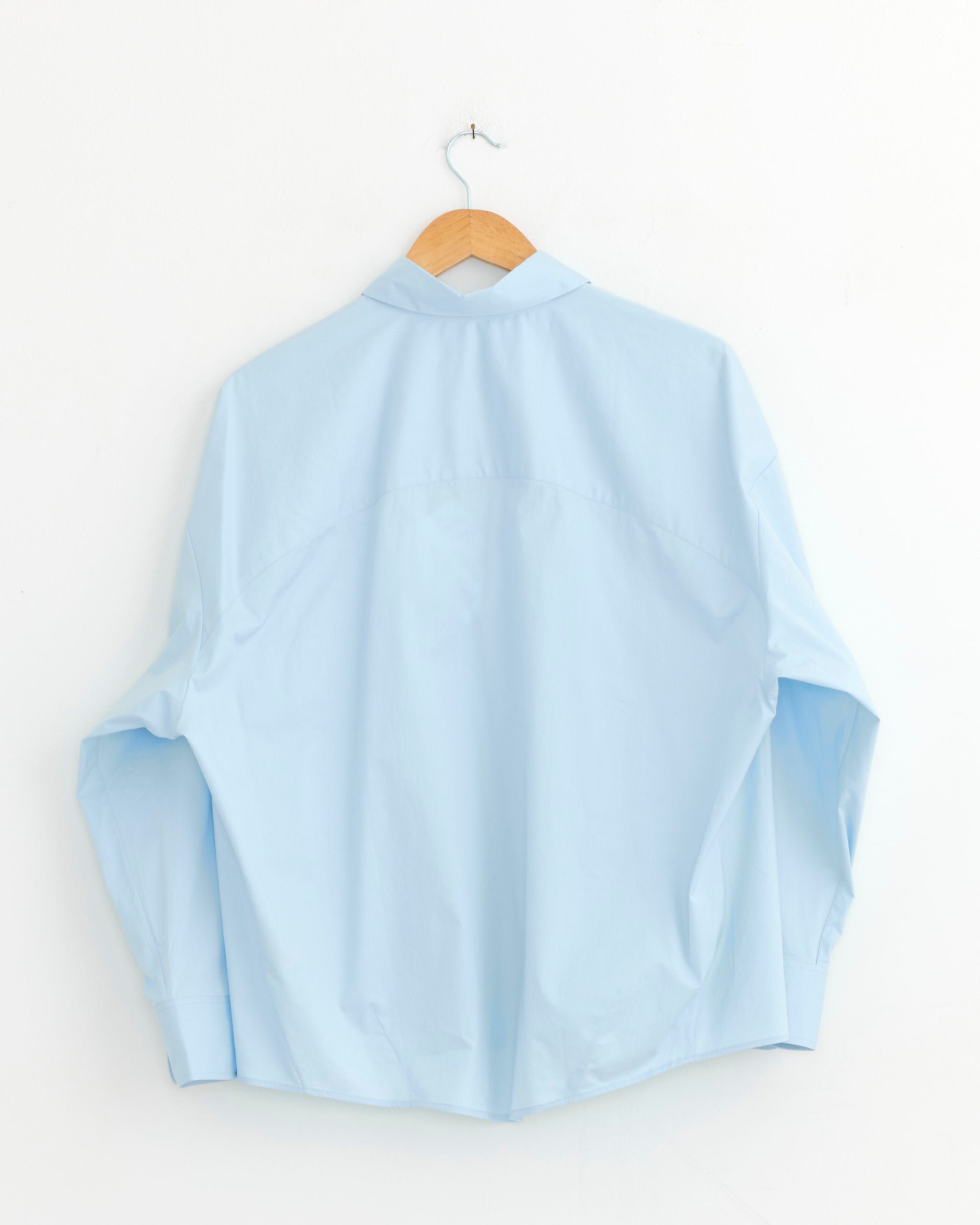The Oversized Shirt in Light Blue by CORDERA features an Italian collar and long sleeves. Displayed from the back, it hangs on a wooden hanger against a plain white wall.