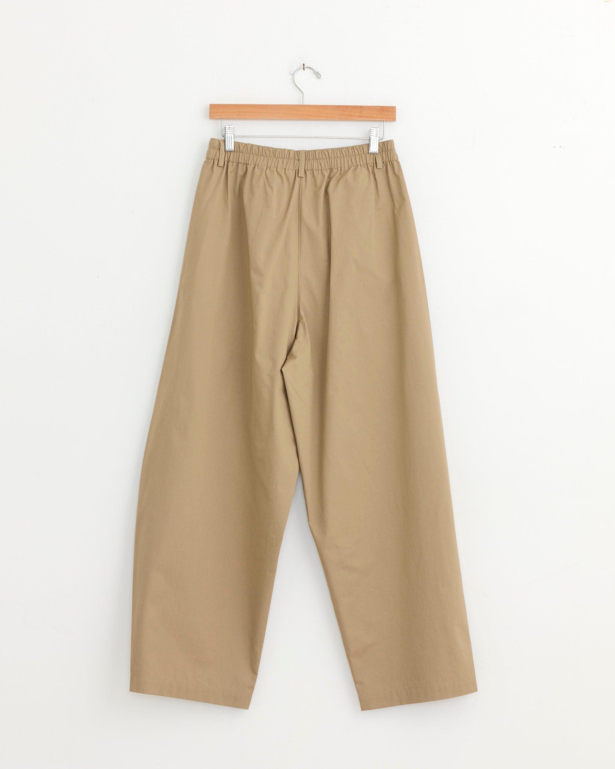 CORDERA Curved Pant in Camel, featuring an oversized silhouette and elastic waistband, hanging on a wooden hanger against a plain white background.