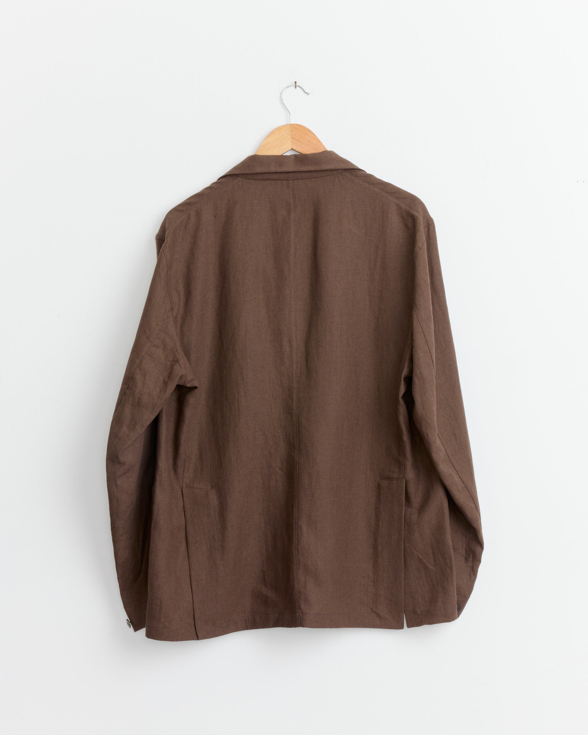 The Single Breasted Shirt Jacket in Chocolate by Stoffa hangs on a wooden hanger against a plain white wall, shown from the back.