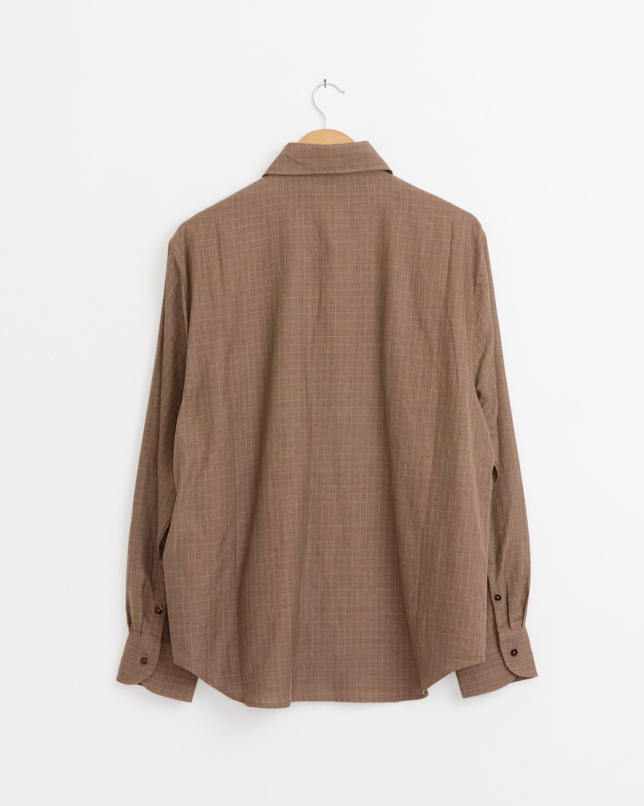 The Spread Collar Shirt in Brown by Stoffa hangs on a wooden hanger against a plain white wall, with the back of the long-sleeve button-up shirt facing forward.