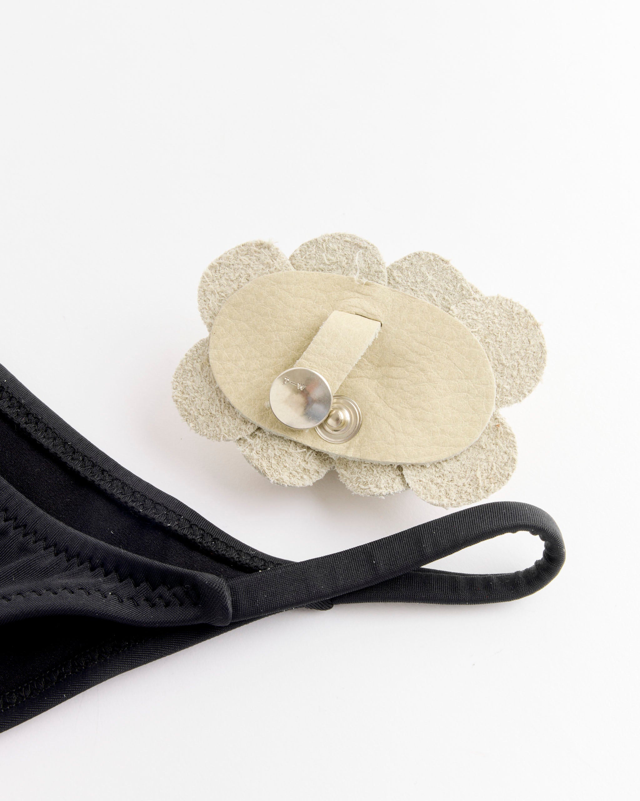 A close-up of a black fabric face mask beside a beige leather flower-shaped clip with metal fastener, echoing the elegant design details of Paloma Wool's Adina Bottom Swimsuit in Black with adjustable side straps.