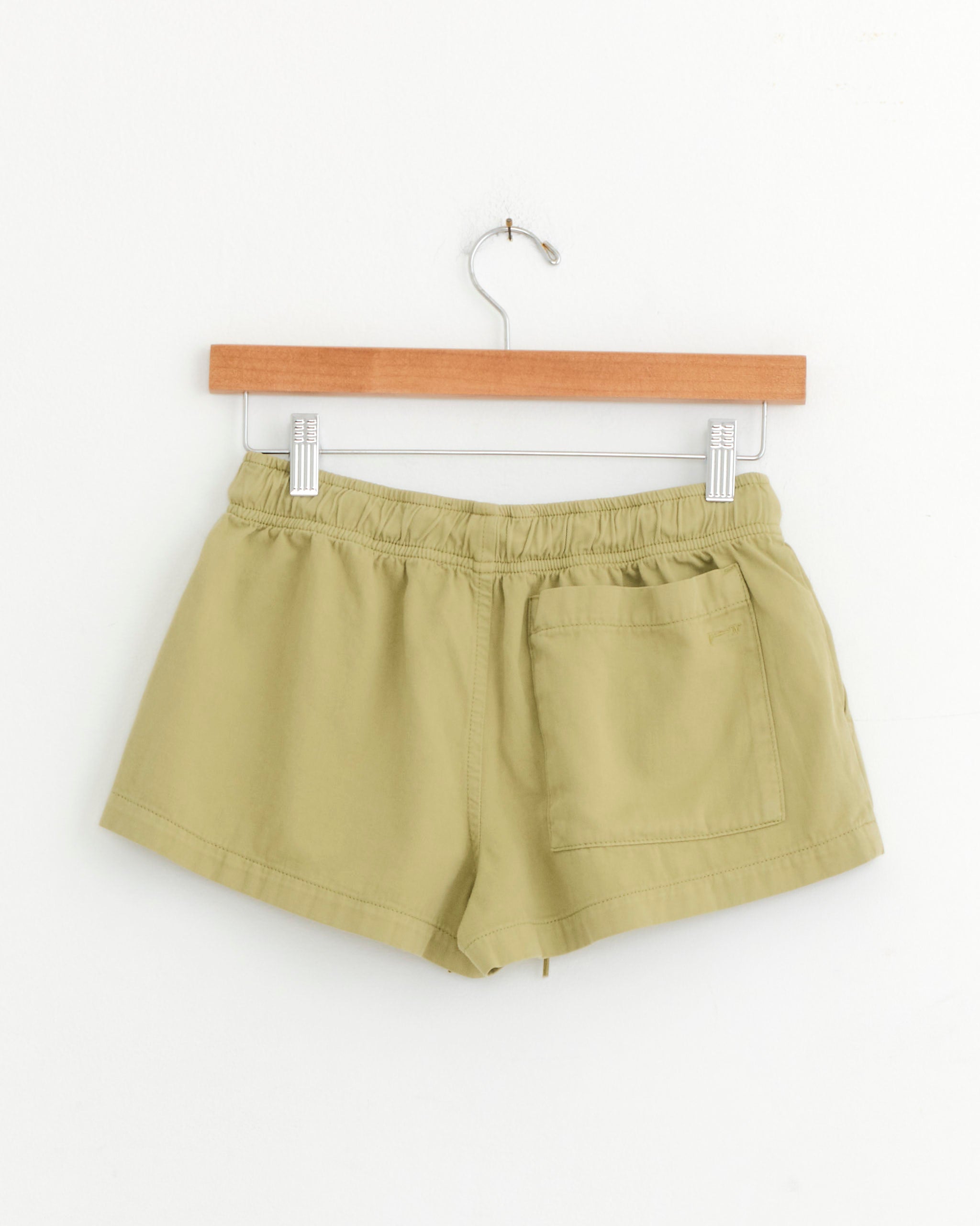 The Paloma Wool Mini Mau Short in Khaki features an elastic waistband and a single back pocket, shown hanging on a wooden hanger against a white background.