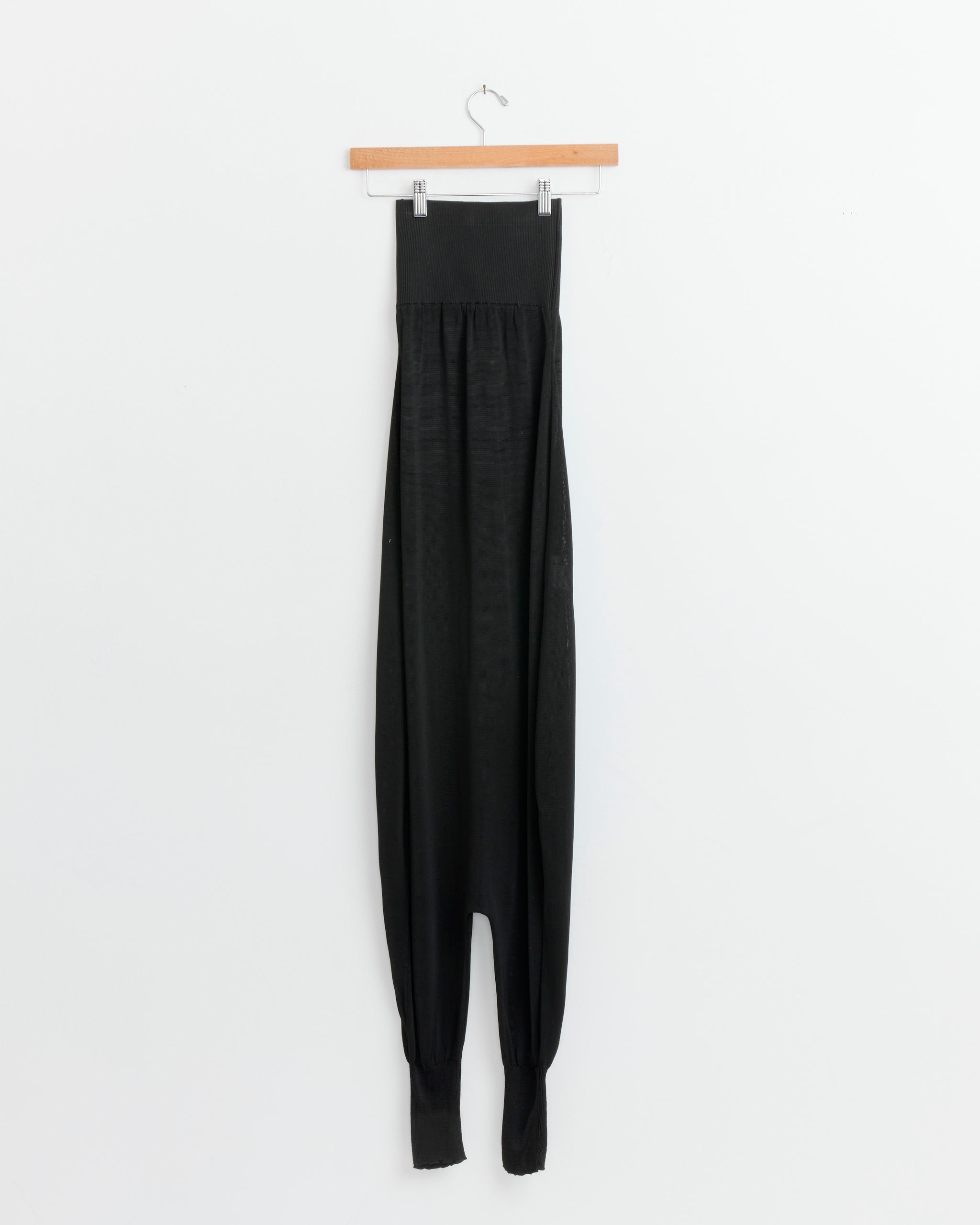 The Gelo Jumpsuit in Black by Paloma Wool, featuring a loose, high-waisted balloon-leg silhouette with tapered legs, hangs from a wooden hanger against a plain white wall.