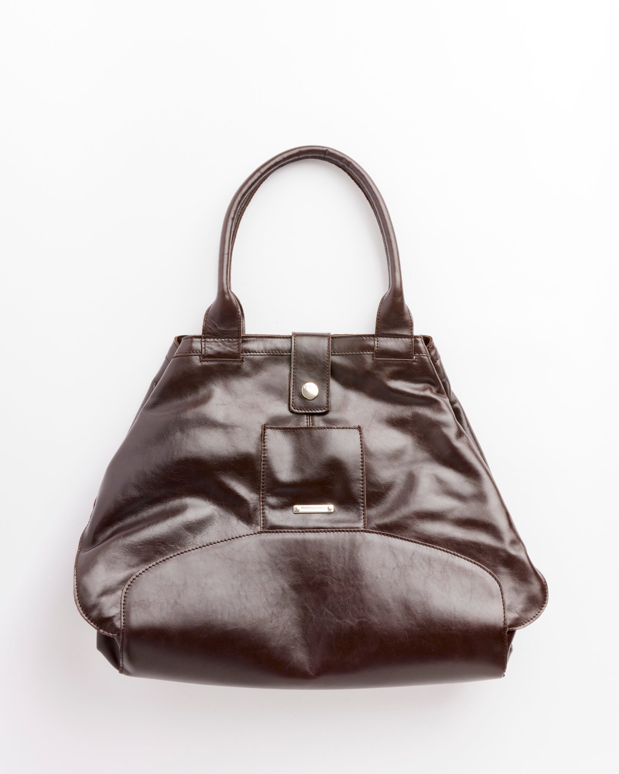 The Paloma Wool Big Guida Bag in Brown is a dark brown 100% leather handbag with two handles, a front snap pocket, and a subtle shine, crafted in España and shown on a white background.