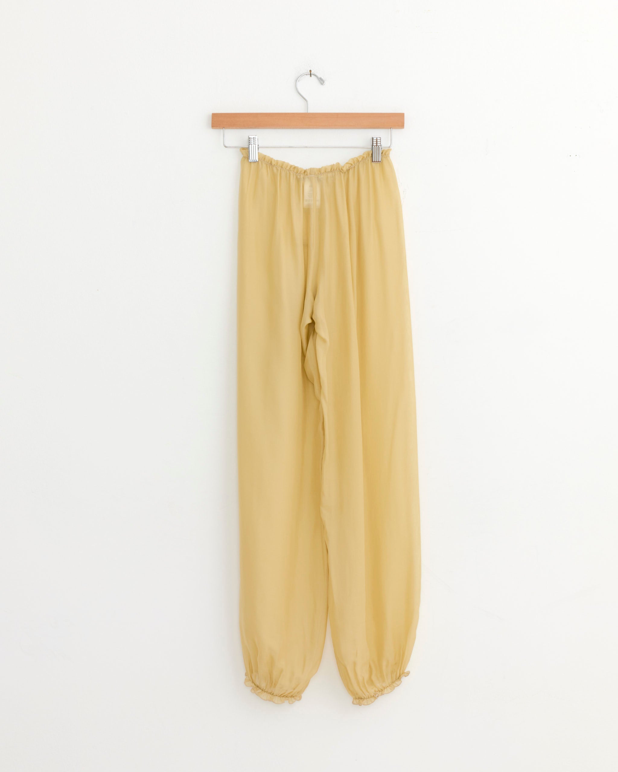 The Weza Pant in Ecru by Paloma Wool features a beige, loose-fitting, lightweight design with elastic waistband and cuffs, displayed on a wooden hanger against a plain white wall.
