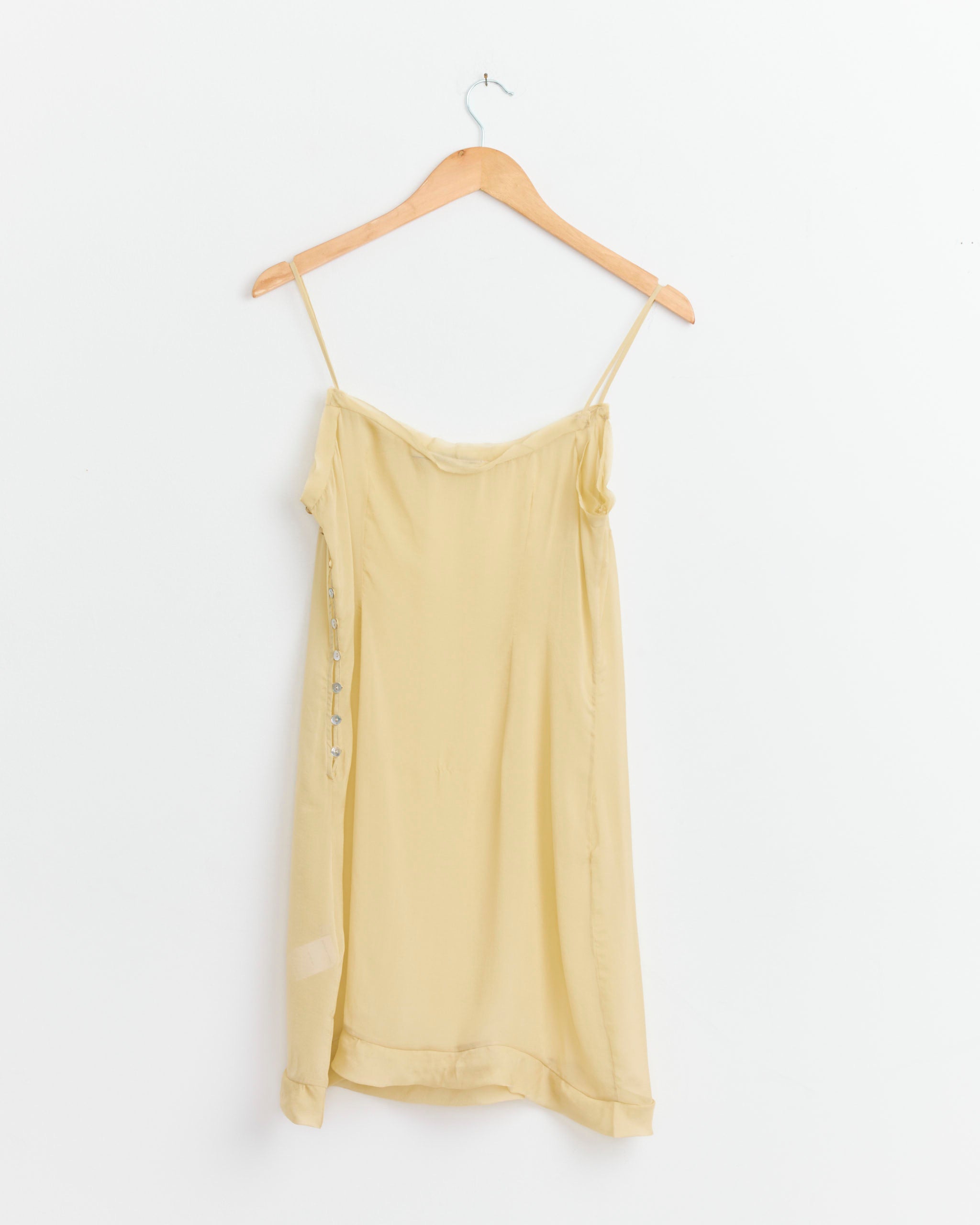 The Paloma Wool Short Enos Dress in Ecru, a light yellow silk slip with thin spaghetti straps and a ruffle hem, hangs on a wooden hanger against a white background. Simple and lightweight, it embodies effortless style.