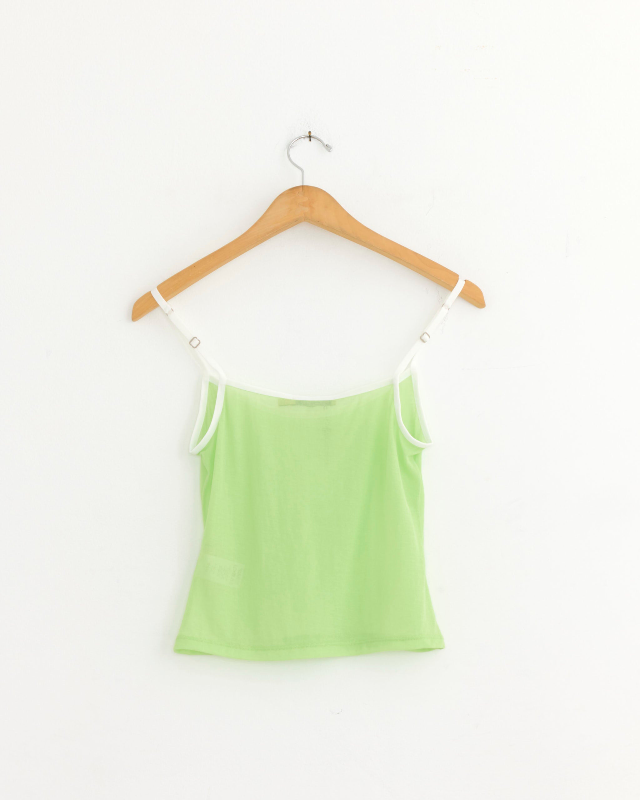 The Paloma Wool Nasem Top in Green, featuring white adjustable spaghetti straps and made from organic cotton, is displayed on a wooden hanger against a plain white wall.
