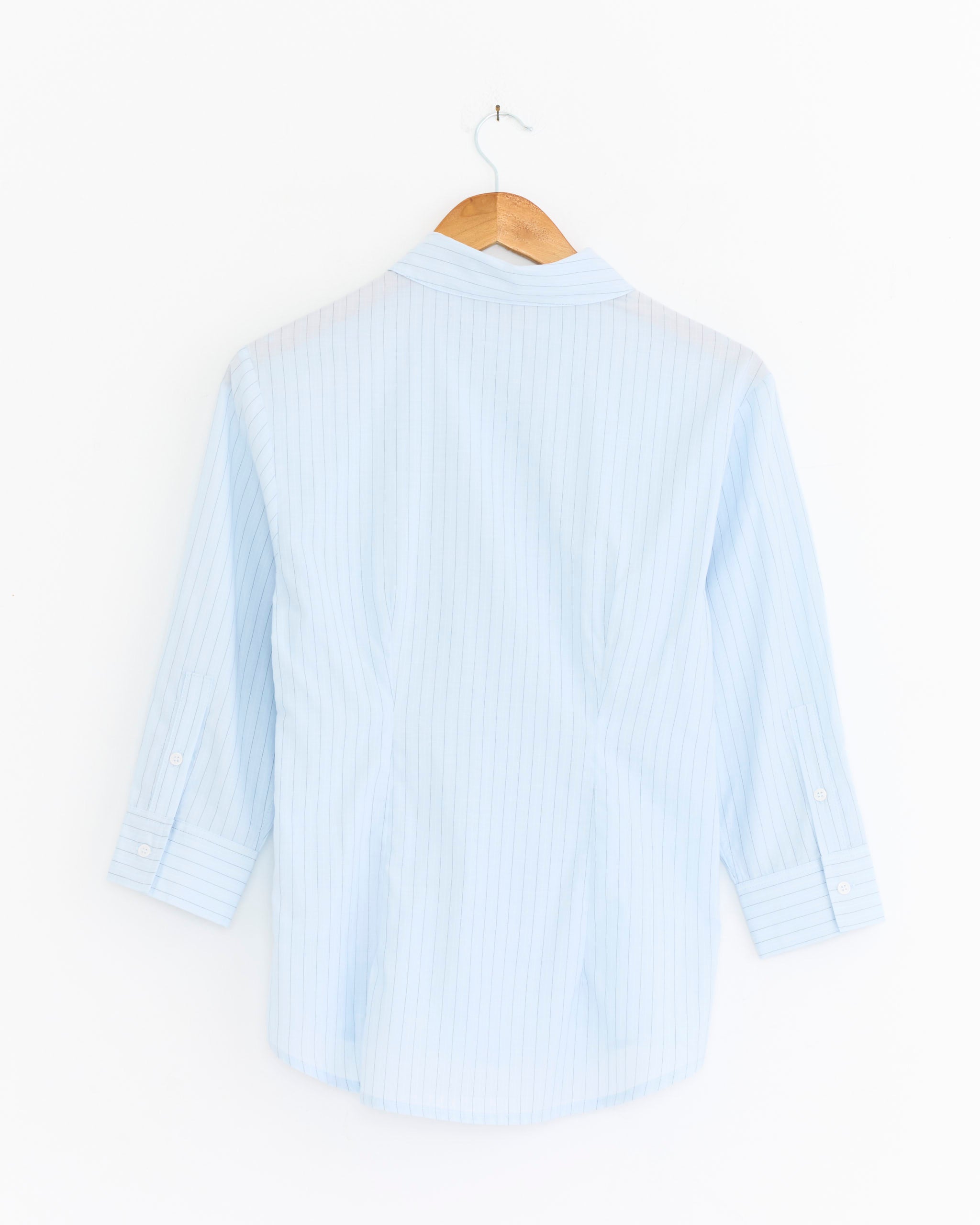 The Bruna Top in Blue by Paloma Wool, made from organic cotton with subtle stripes, has long sleeves and button-up detailing. It hangs on a wooden hanger with the back facing the viewer against a white background.