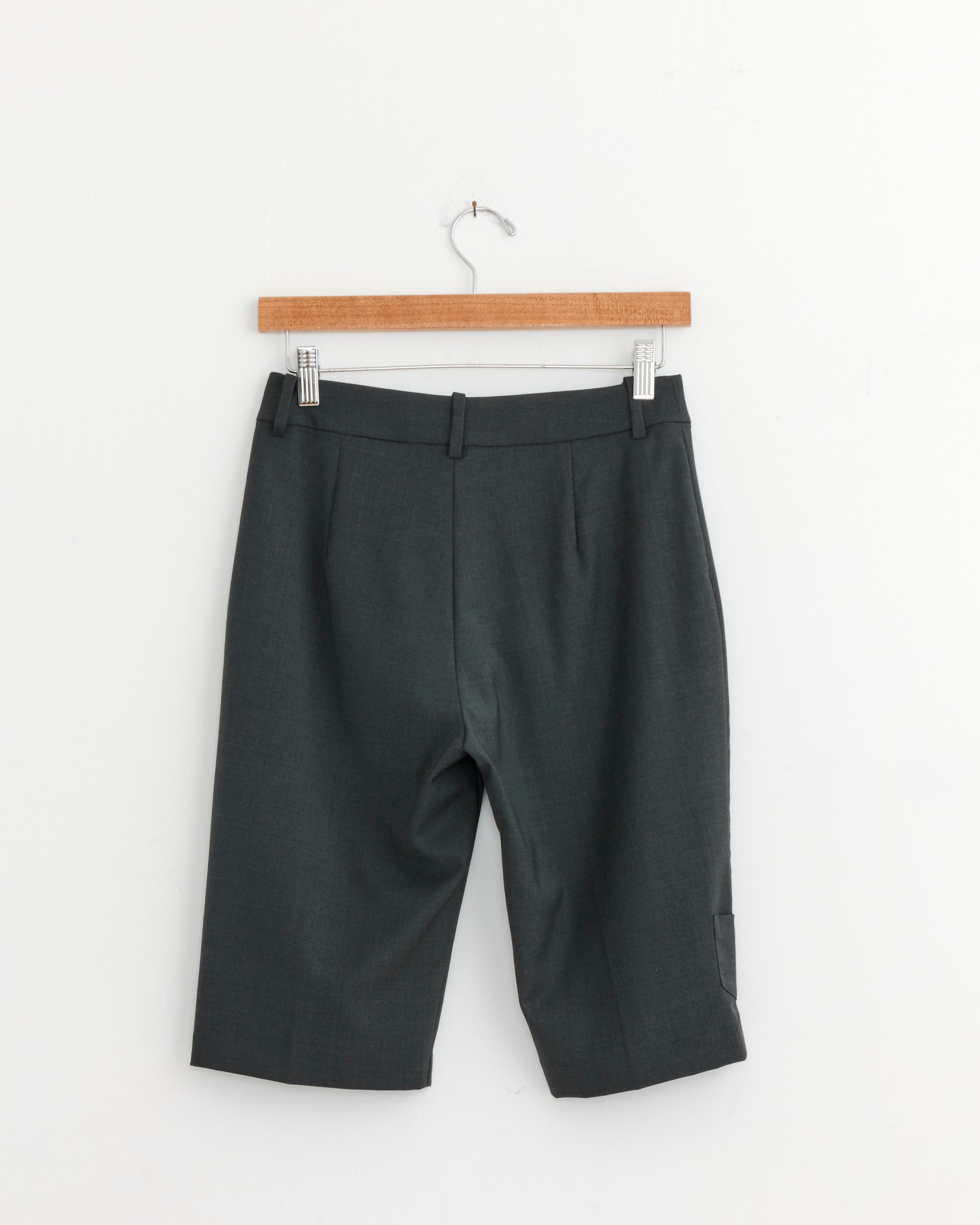 The Marisa Capri in Grey by Paloma Wool, made from recycled polyester with belt loops and a knee-length cut, hangs on a wooden hanger against a plain white wall.