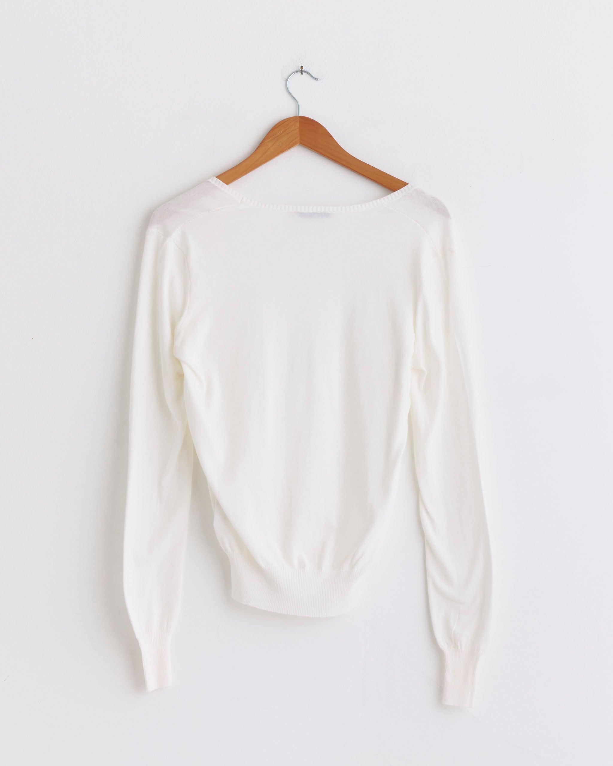 The Paloma Wool Omina Sweater in Off White hangs on a wooden hanger against a plain white wall, highlighting its timeless long-sleeved style.