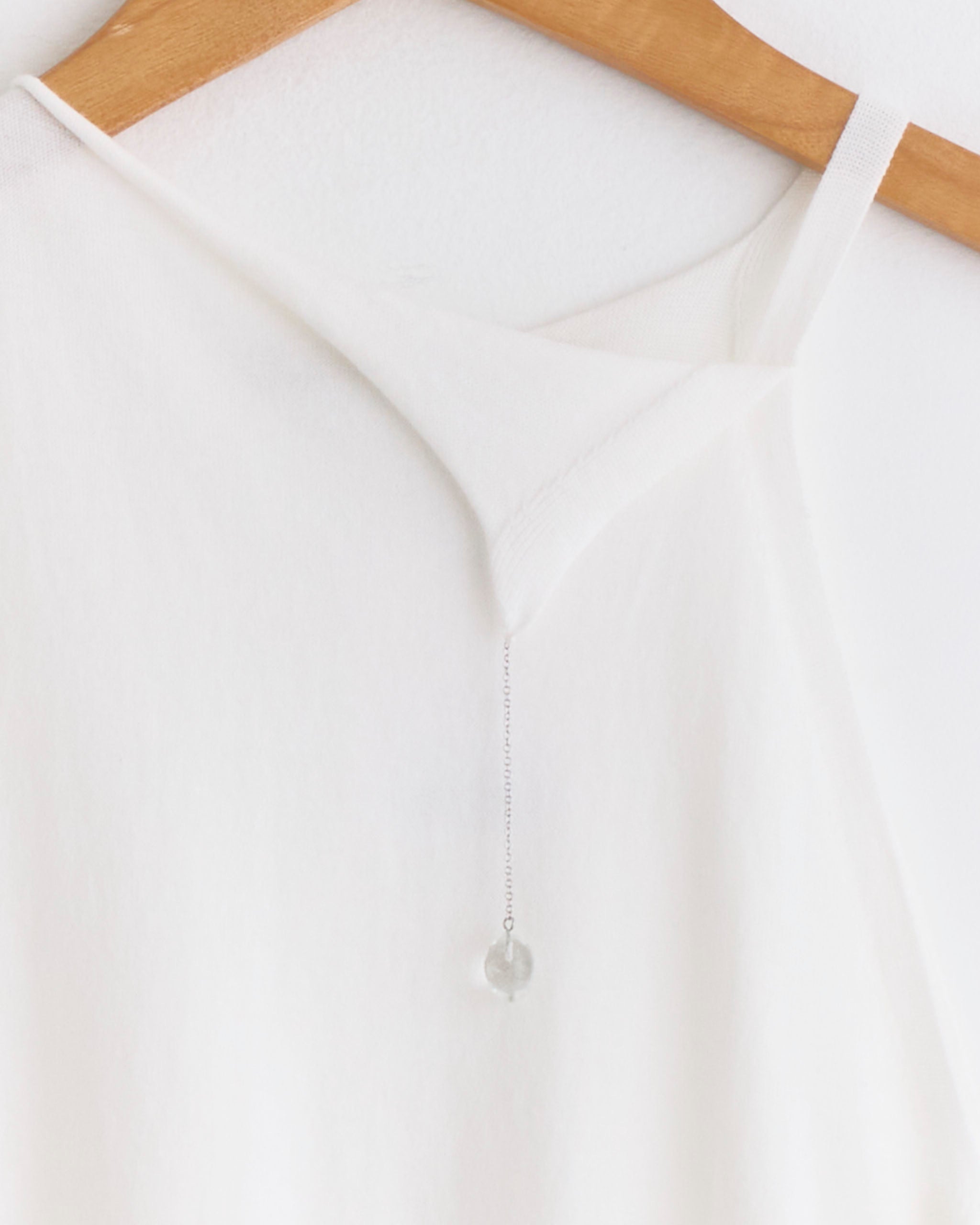 The Lani Tank in Off White by Paloma Wool, a sleeveless knit top, hangs on a wooden hanger against a white wall. A delicate silver chain with a clear round pendant drapes from its neckline.