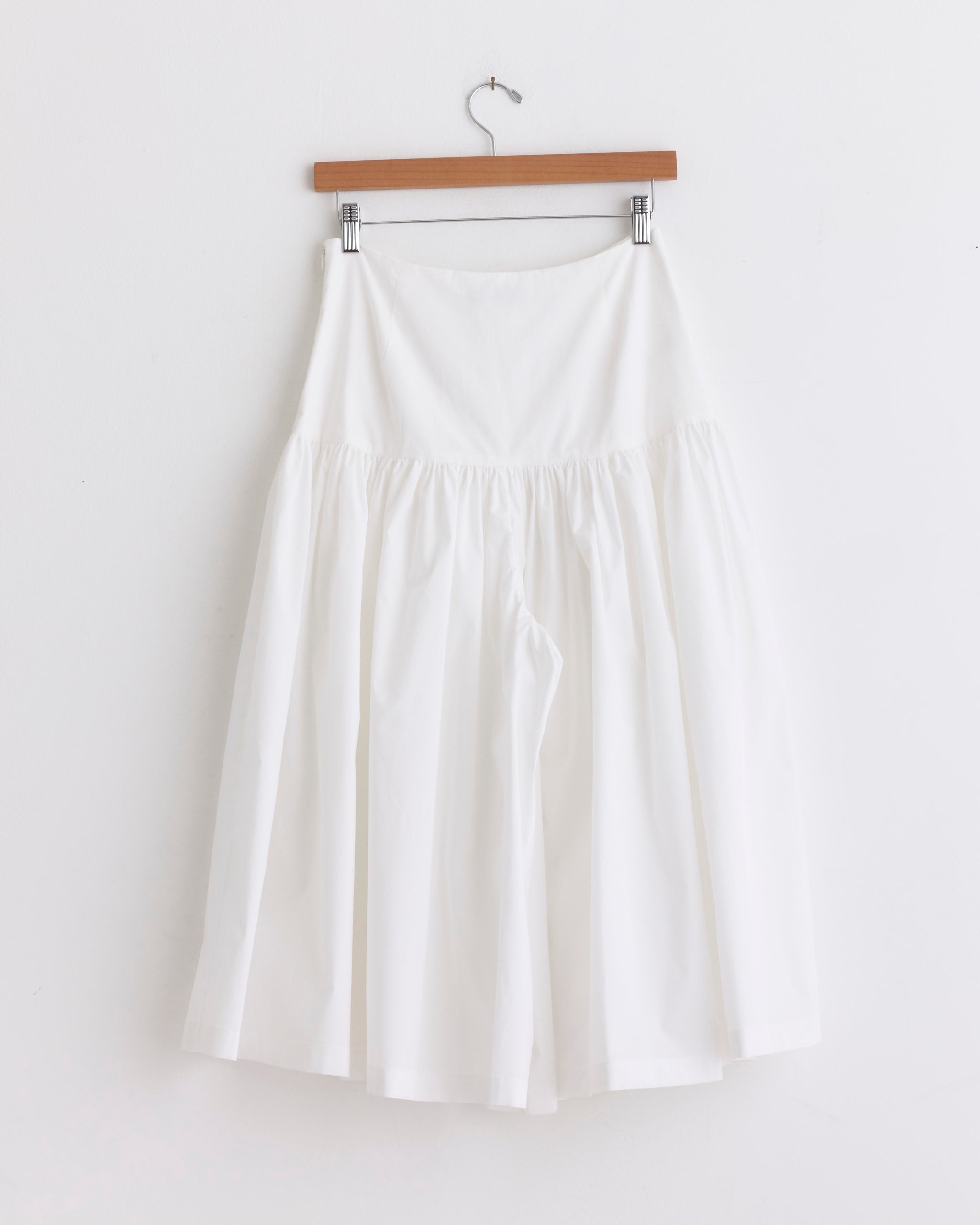 The Amomento Shirring Culotte Pant in White, featuring a wide fit and skirt-like silhouette, is displayed on a wooden hanger against a plain white wall.