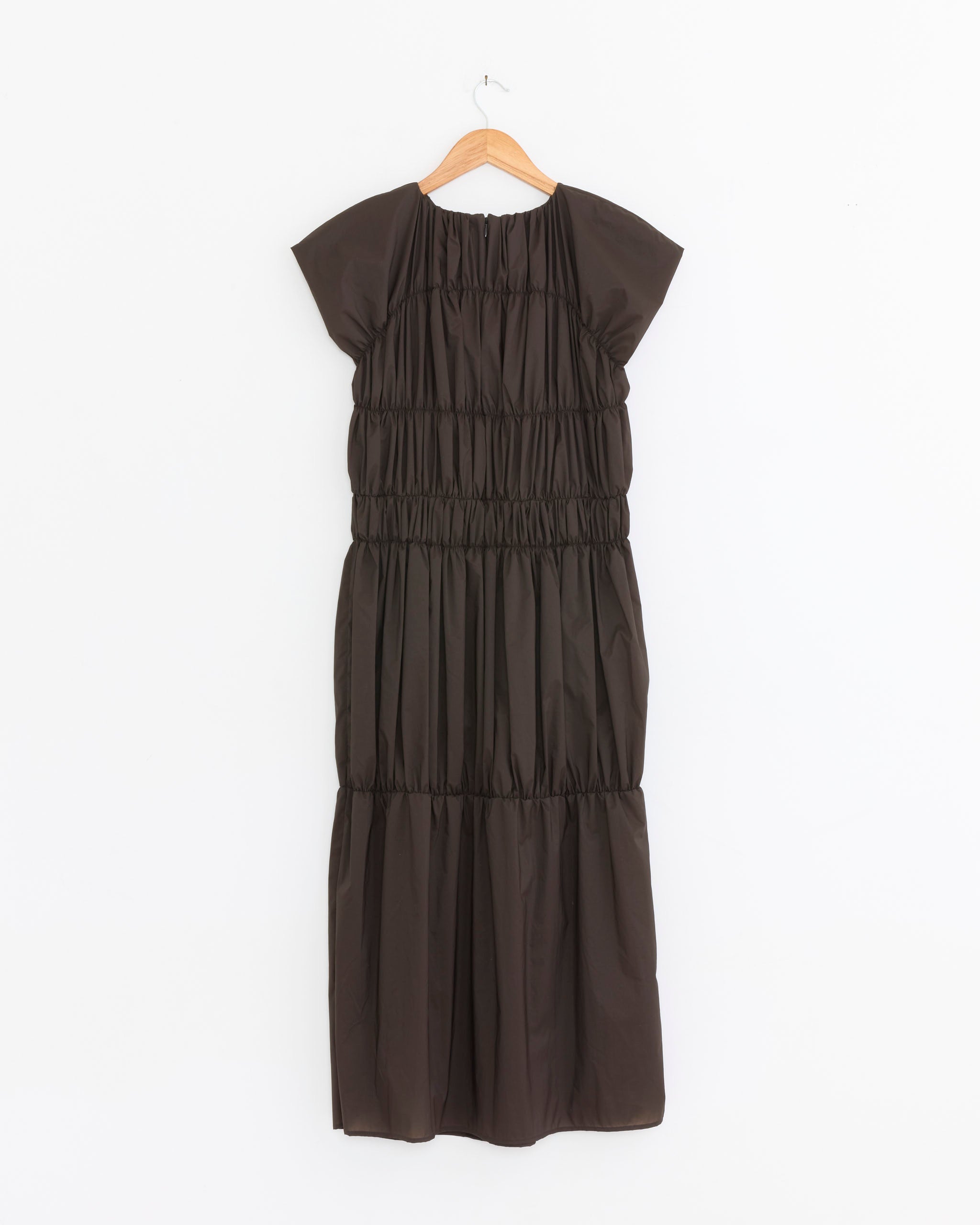 The Amomento Nylon Shirring Dress in Brown is a short-sleeved, loose-fit dress with smocked shirring detail, made from breathable nylon, shown on a wooden hanger against a white background.