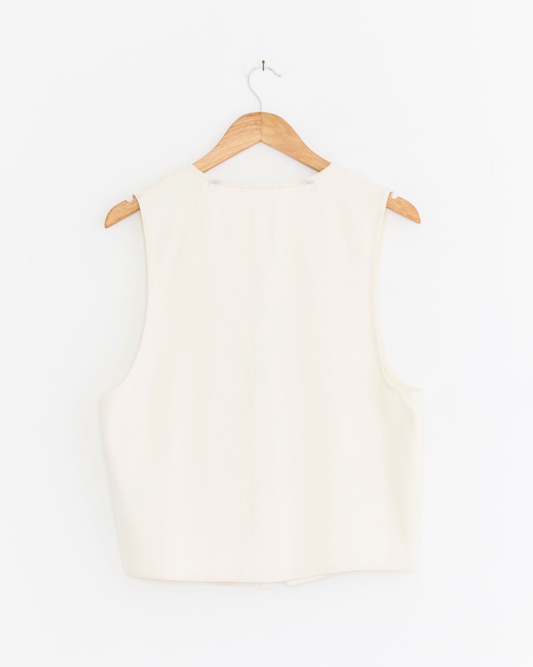 The Comoli Silk Nep Vest in Ecru, made in Japan, hangs sleeveless on a wooden hanger against a plain white wall, displayed from the back.