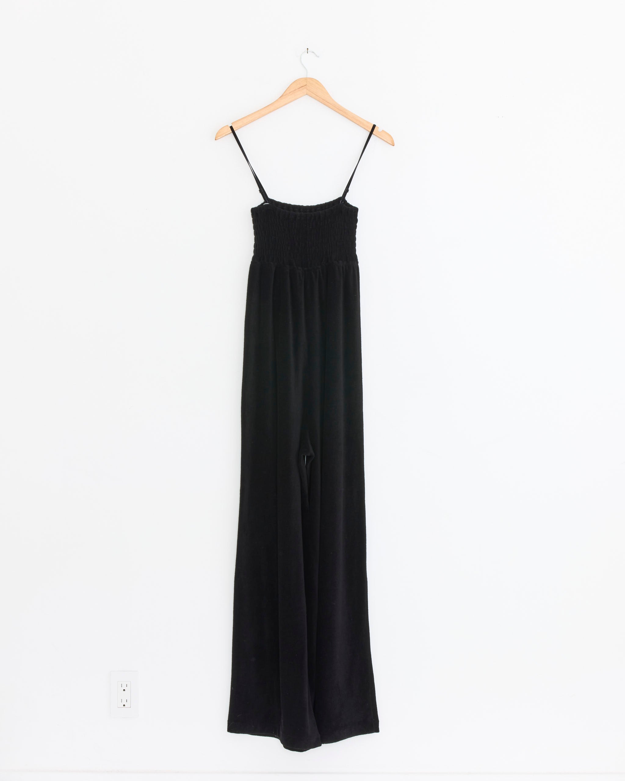 The Comoli Pile Bare Top Salopette Pant in Black, a sleeveless wide-leg jumpsuit with thin spaghetti straps, hangs on a wooden hanger against a plain white wall.