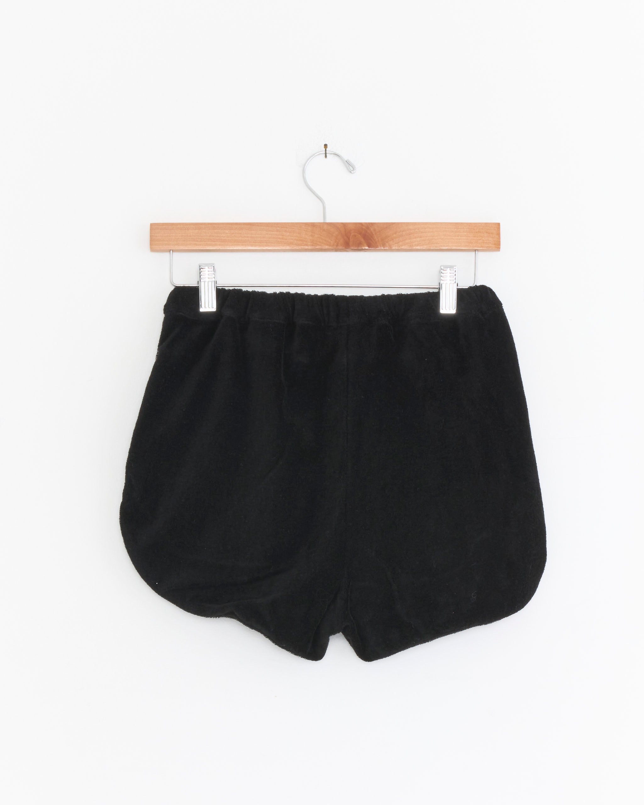 The Comoli Pile Shorts in Black, made from lightweight, breathable cotton terry with an elastic waistband, hang on a wooden hanger against a plain white background.