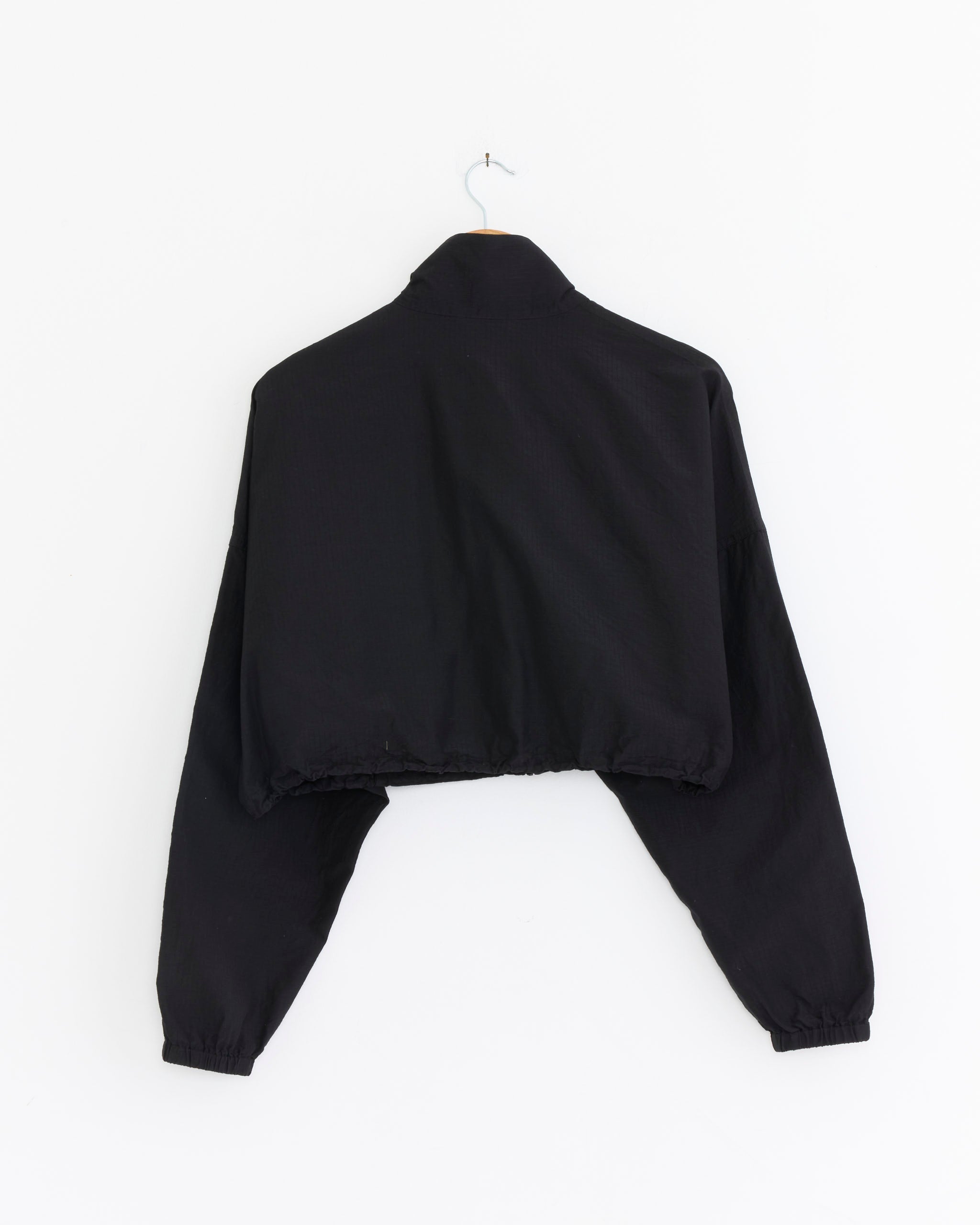 The Comoli Ripstop Short Jacket in Black, featuring long sleeves and a cropped design, is displayed from the back on a simple hanger against a plain white background.