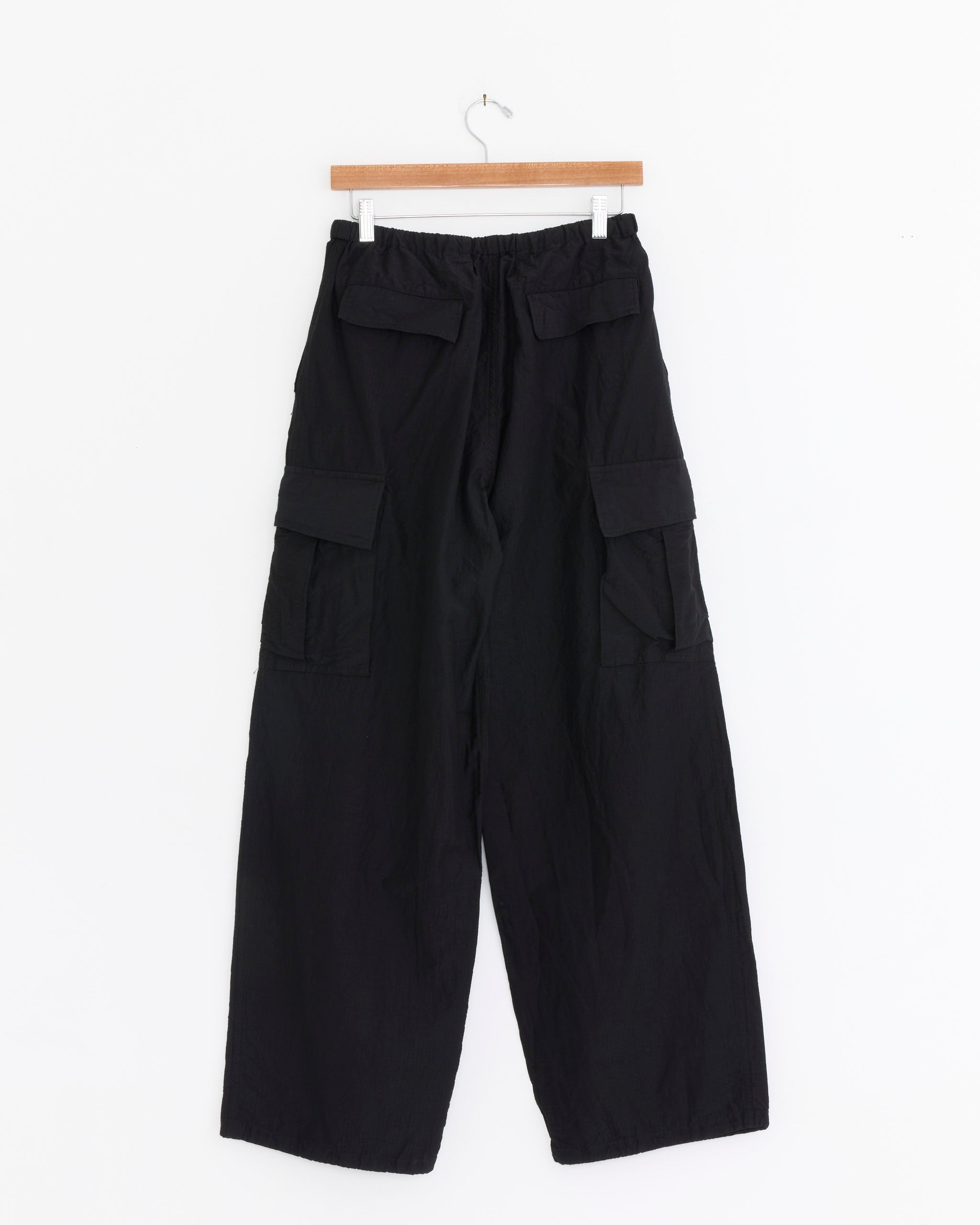 Comoli Ripstop 6-Pocket Pant in Black, featuring an elastic waistband, wide legs, and multiple pockets, is displayed on a wooden hanger against a plain white background.