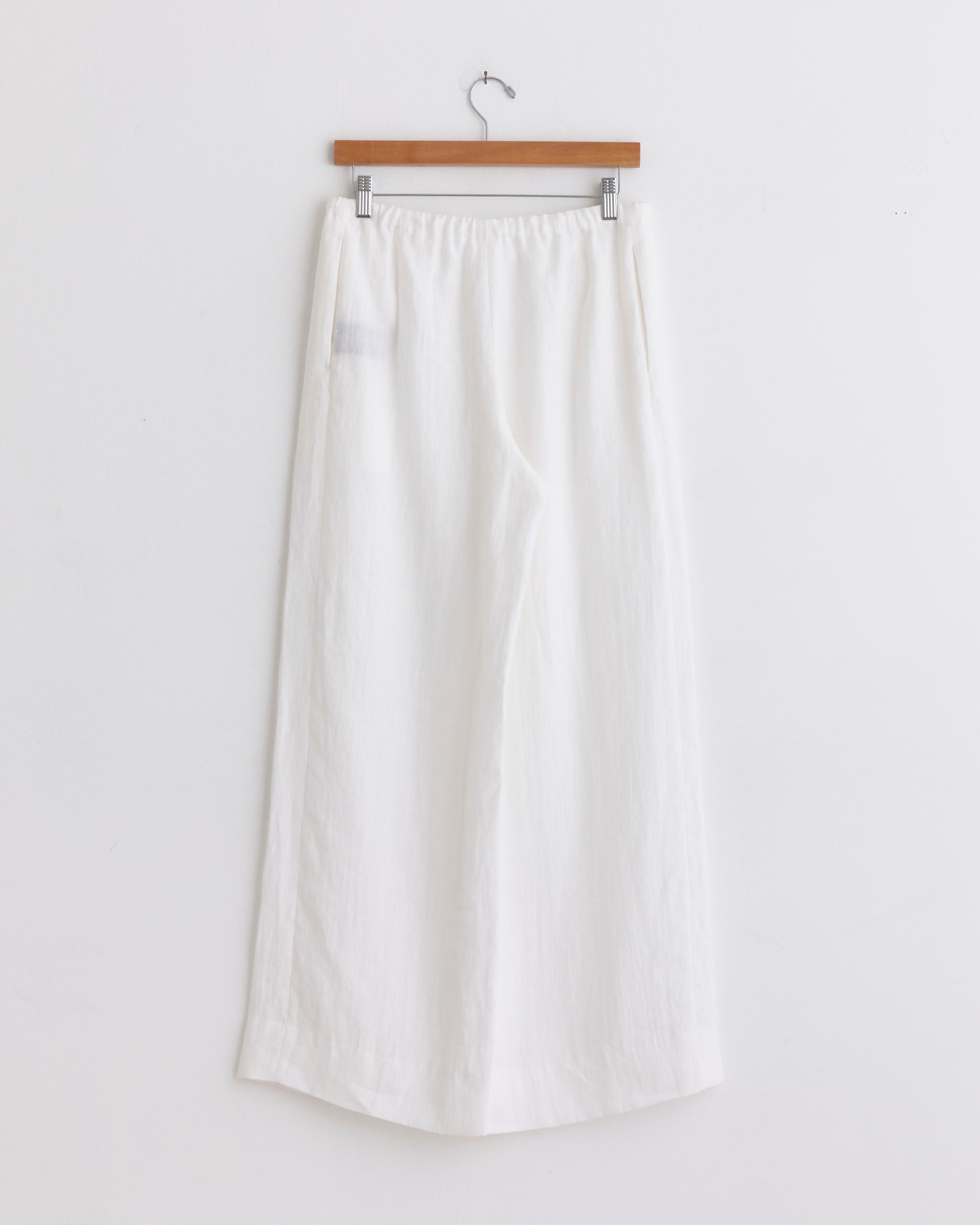 The Canapa Wide Pant in White by Comoli hangs on a wooden hanger against a white wall. Made in Japan from lightweight hemp, these pants feature an elastic waistband and relaxed fit, embodying Comoli's minimalist style.